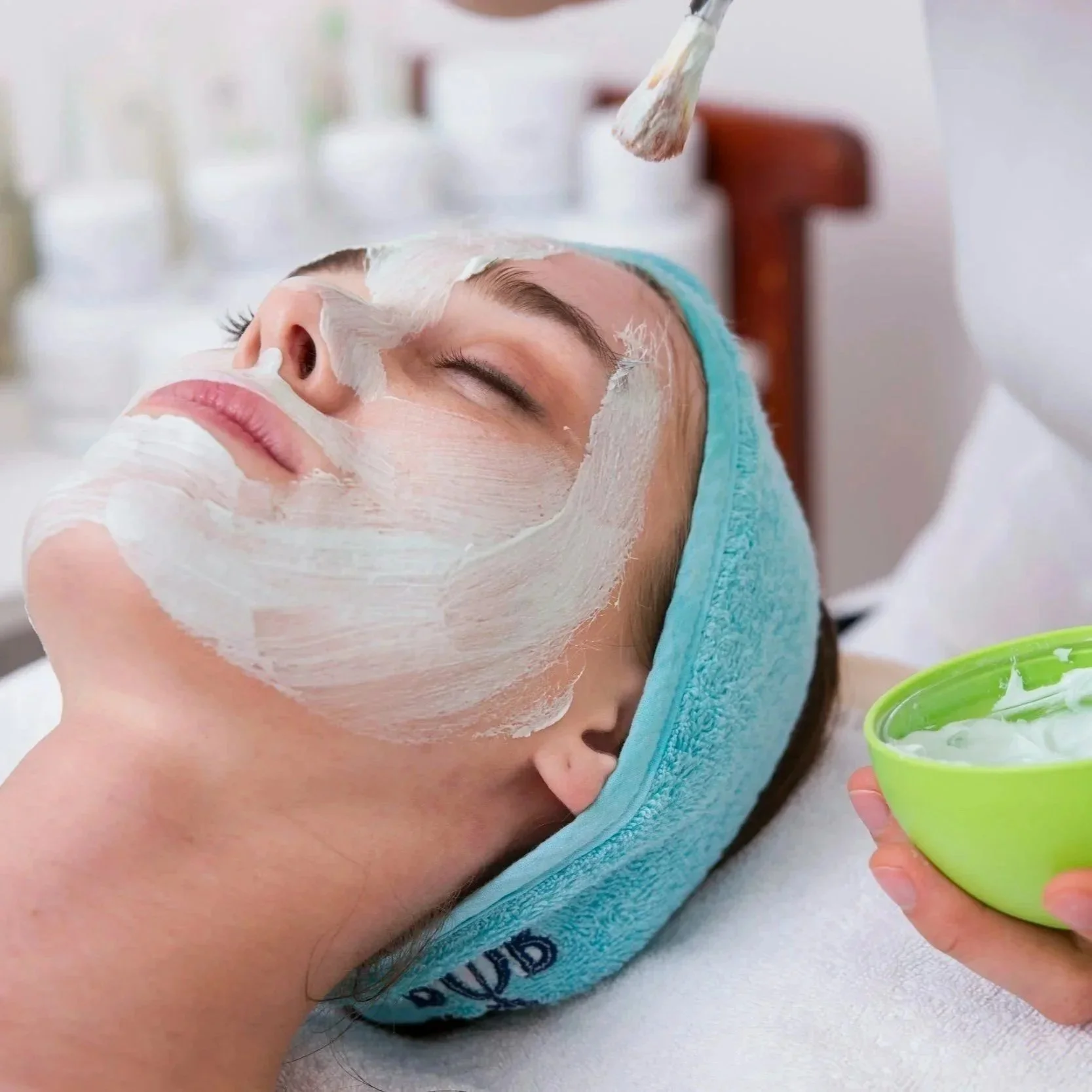 Woman receiving a facial treatment with a white facial mask applied, lying down with a blue towel wrapped around her head, and holding a green bowl of cream or moisturizer.