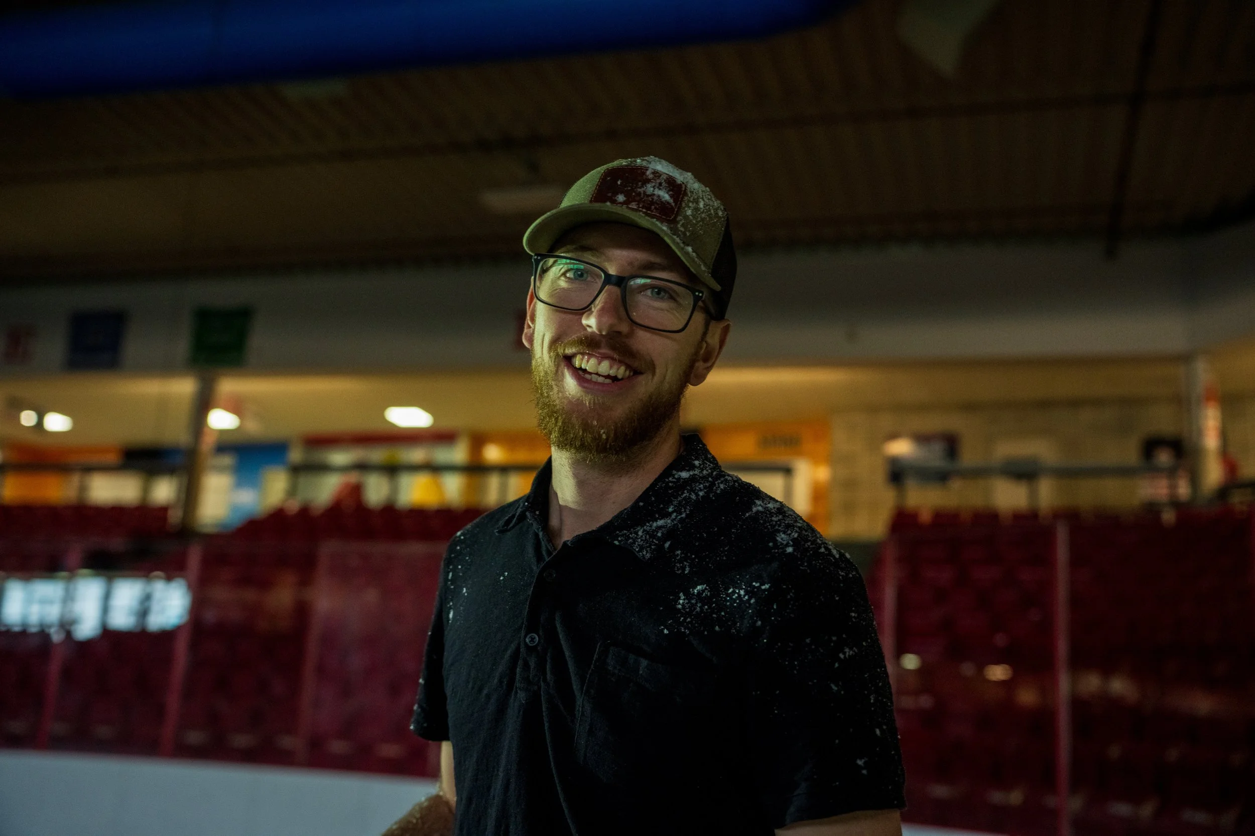 A smiling man with glasses, a beard, and a camo hat stands indoors with a dark background and red fencing, wearing a black shirt covered with white powder or dust.