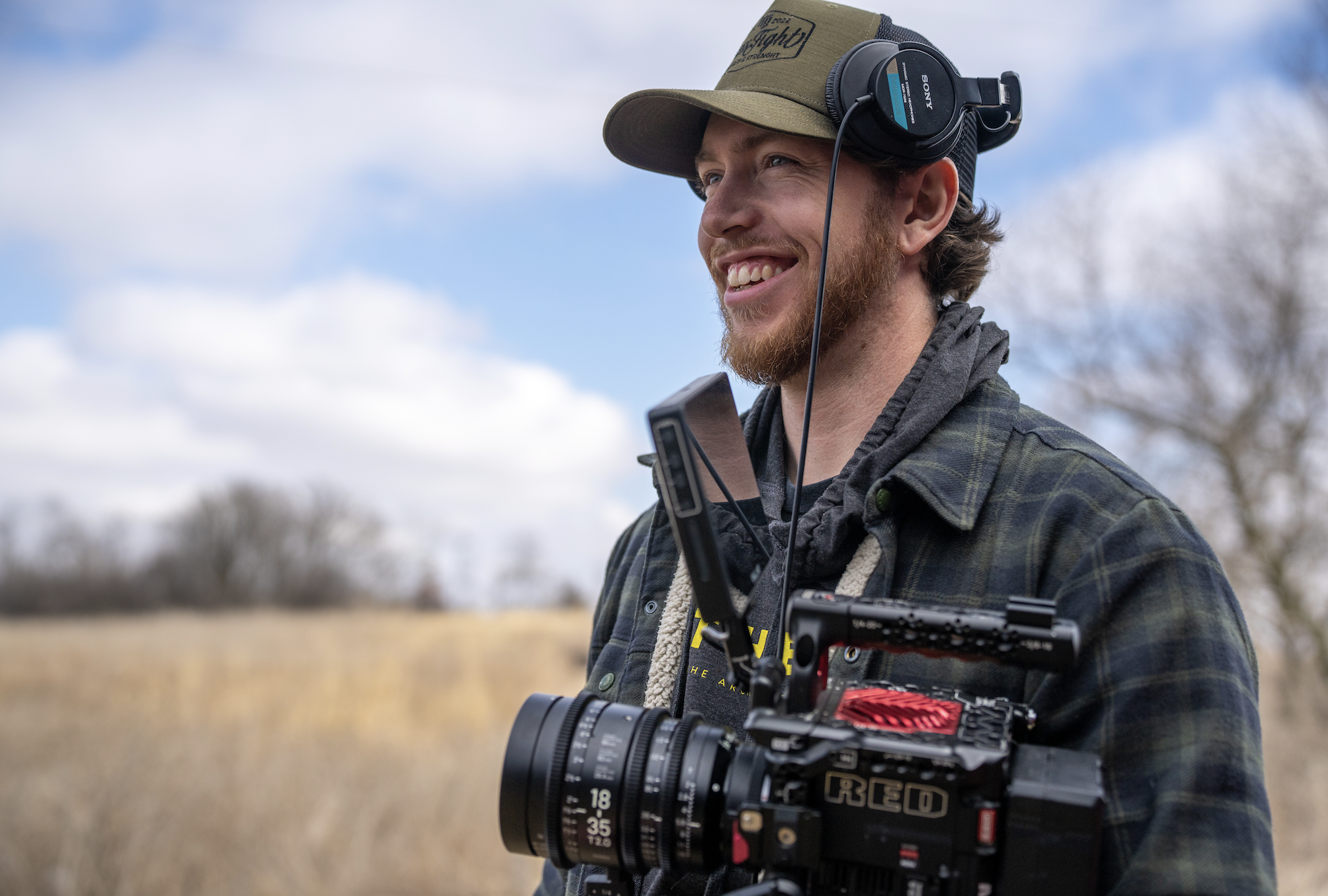 A man outdoors in a field with trees and a partly cloudy sky, wearing a cap, headphones, and a plaid jacket, holding a professional video camera.