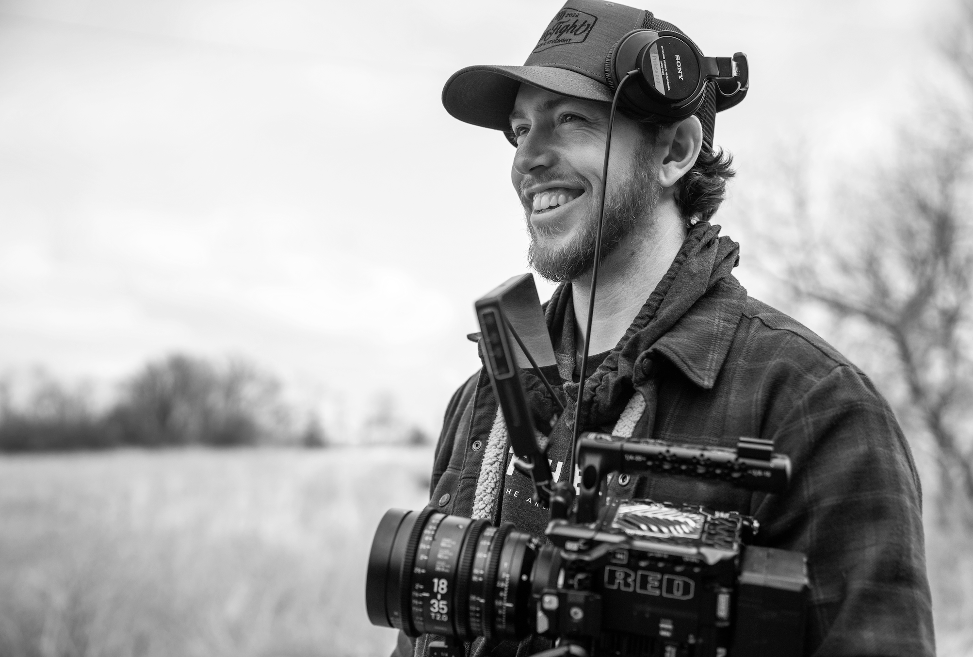 A man with a beard wearing a cap, headphones, and a plaid jacket, smiling outdoors while carrying a film camera mounted on his shoulder.