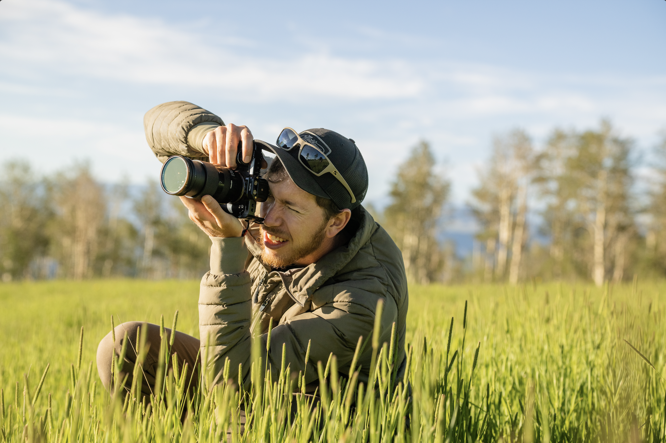 Man taking a photograph with a camera in a grassy field on a sunny day.