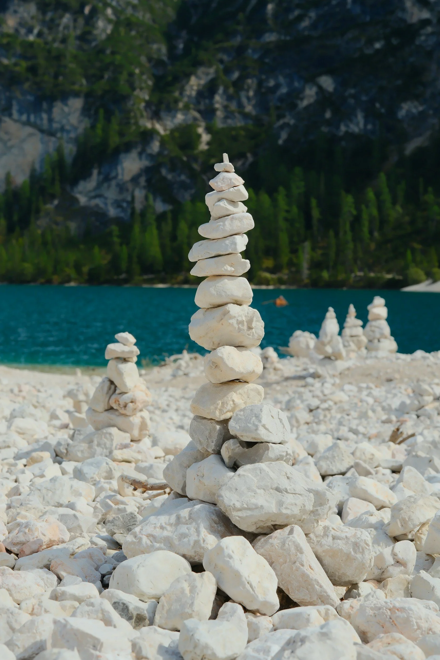 Stacked white rocks on a rocky beach with a lake, forest, and mountains in the background.