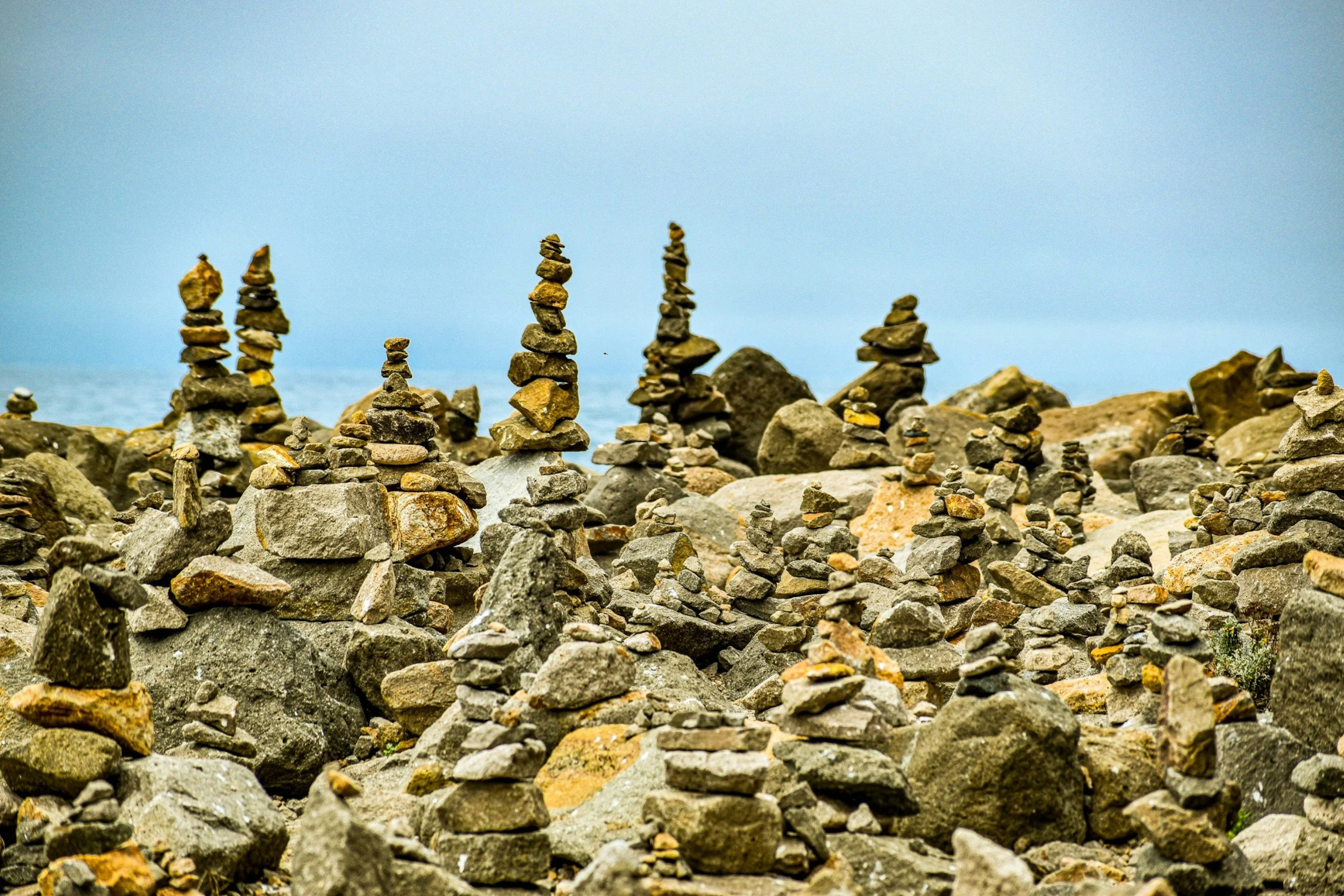 A family of small and large rocks with numerous stacked stone cairns, or waymarks, on a rocky shore near the sea, under a cloudy sky.
