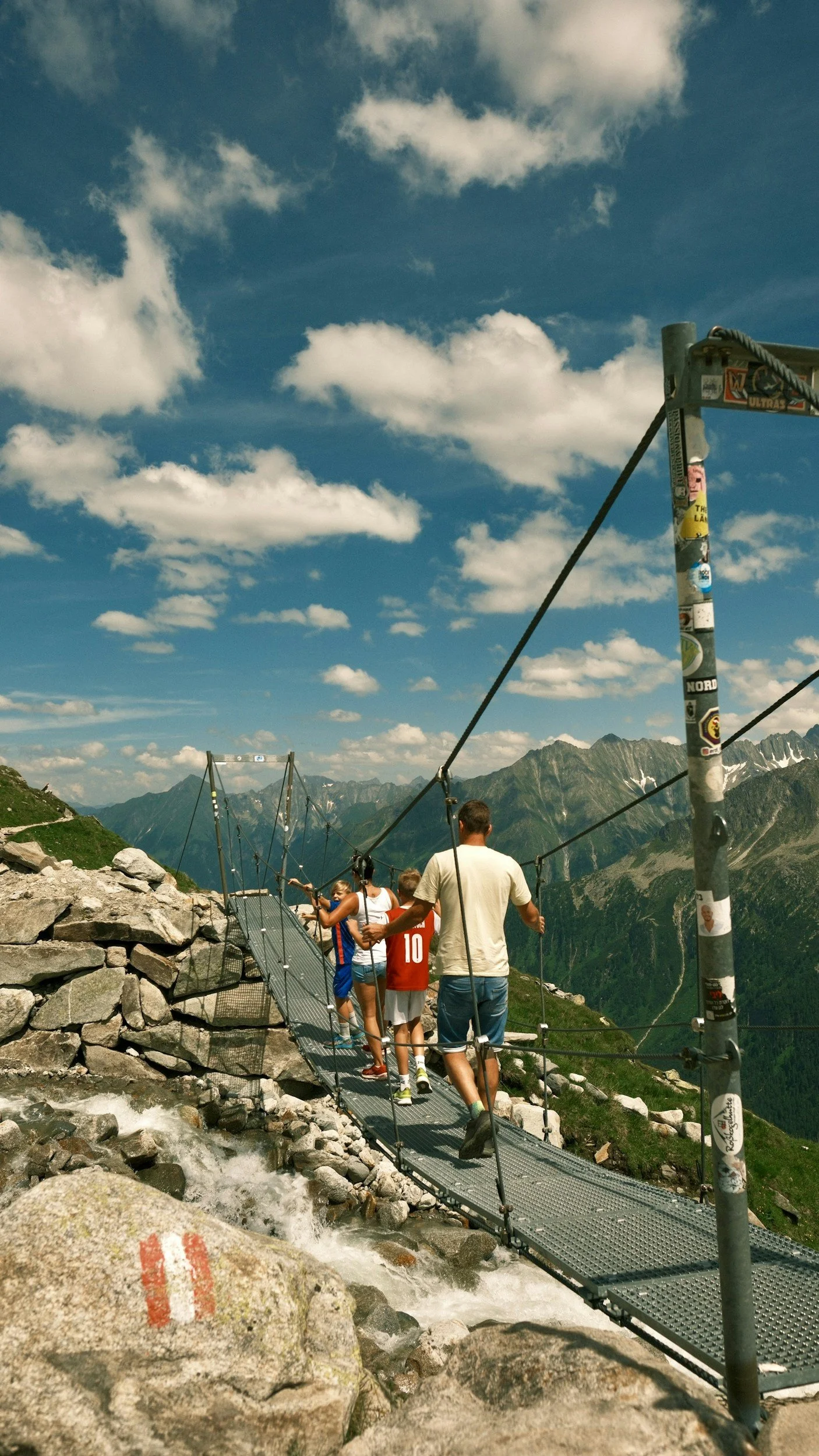 A family crossing a suspension bridge high in the mountains on a clear, partly cloudy day.