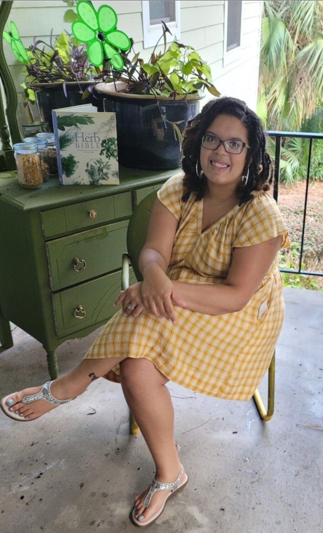 A woman with glasses and curly hair sitting on a green chair on a porch, wearing a yellow checkered dress and sandals. There is a green dresser with a book titled 'The Herb Bible', a large potted plant with green and purple leaves, and glass jars on the dresser. The porch has a railing, and greenery can be seen outside.