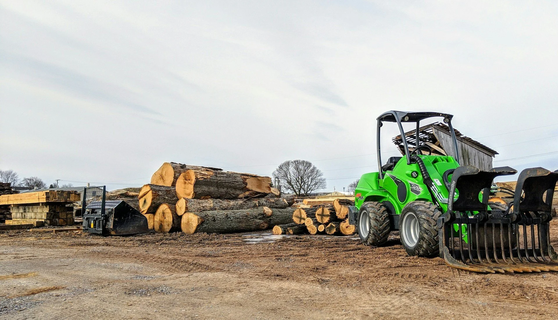 A bright green compact loader with a black fork attachment parked on a dirt lot next to a large pile of cut logs, with an old wooden shed in the background and a cloudy sky overhead.