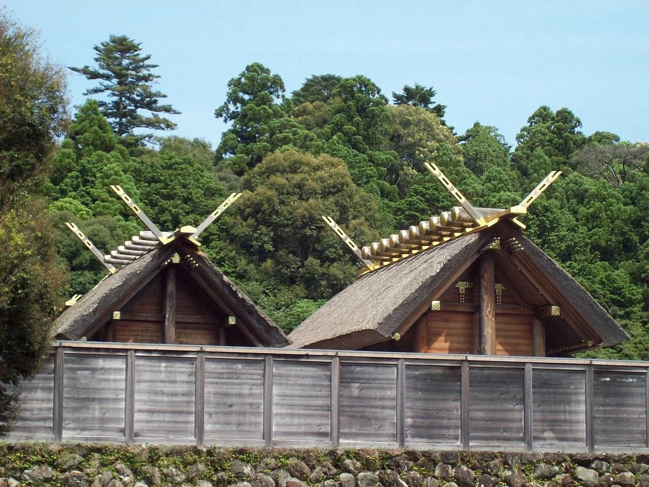 Traditional Japanese wooden buildings with thatched roofs, surrounded by a wooden fence and lush green trees in the background.