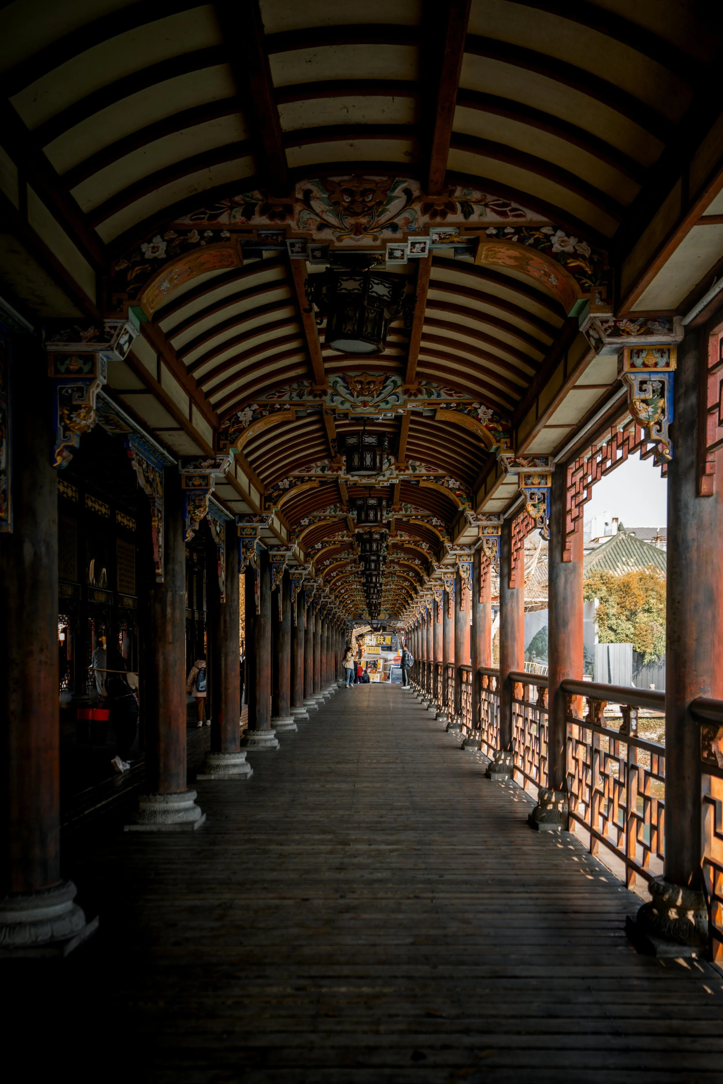 A long covered wooden walkway with ornate painted beams and lanterns, supported by wooden pillars, leading to an area with small shops or stalls at the end.