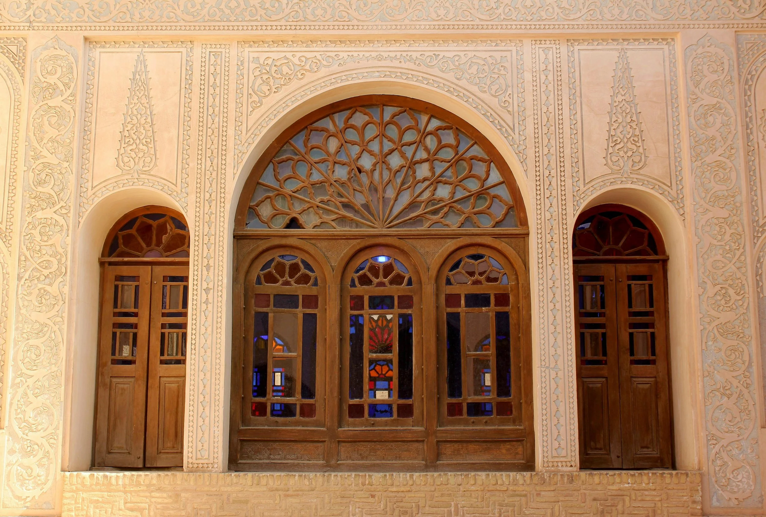 A decorative beige wall with intricate carvings and three arched windows with stained glass. The central window features a fan-shaped wooden lattice design, while the side windows are framed with wood and have geometric stained glass panels.