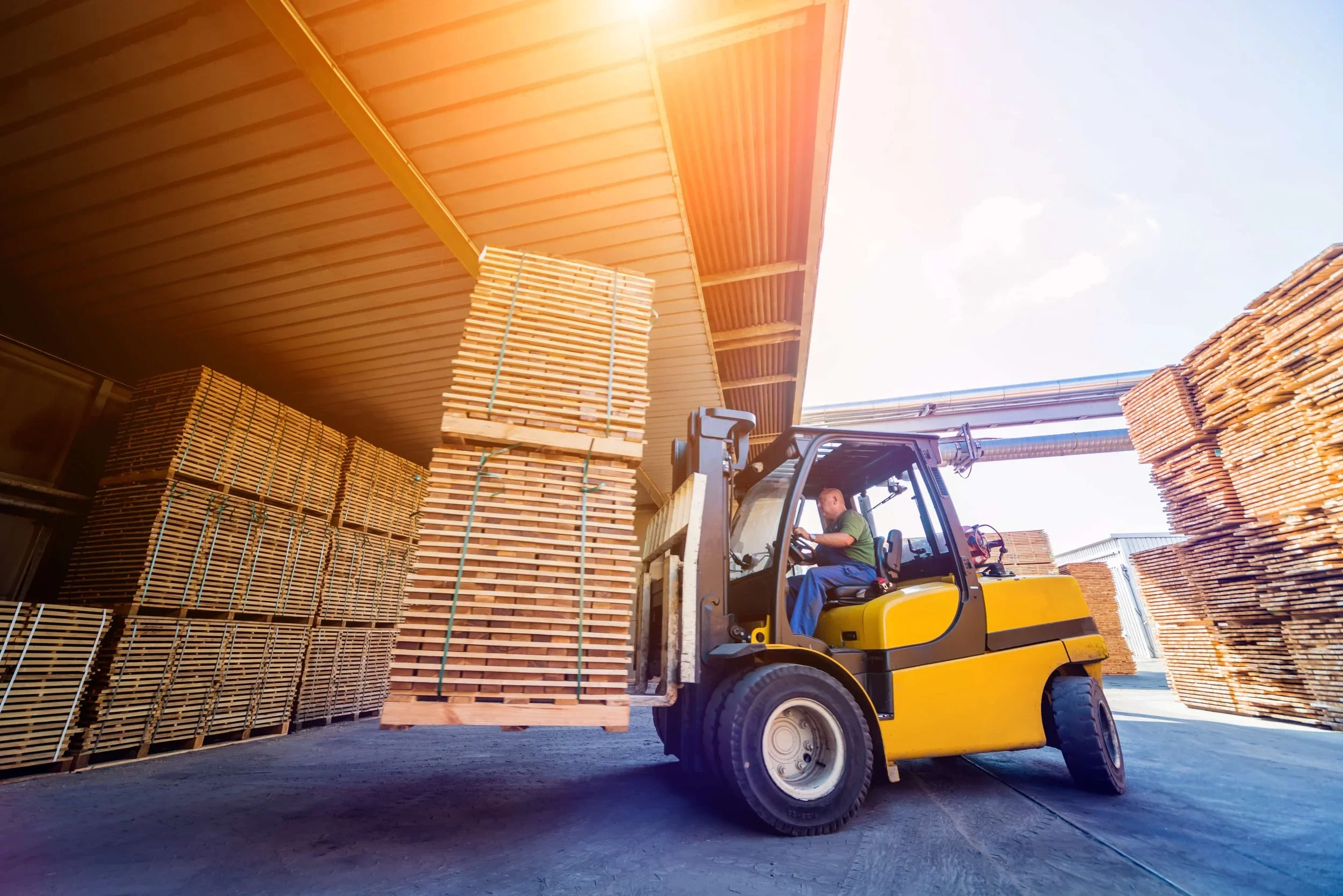 A worker operating a forklift lifting wooden pallets in a warehouse with pallets stacked on both sides and the roof overhead