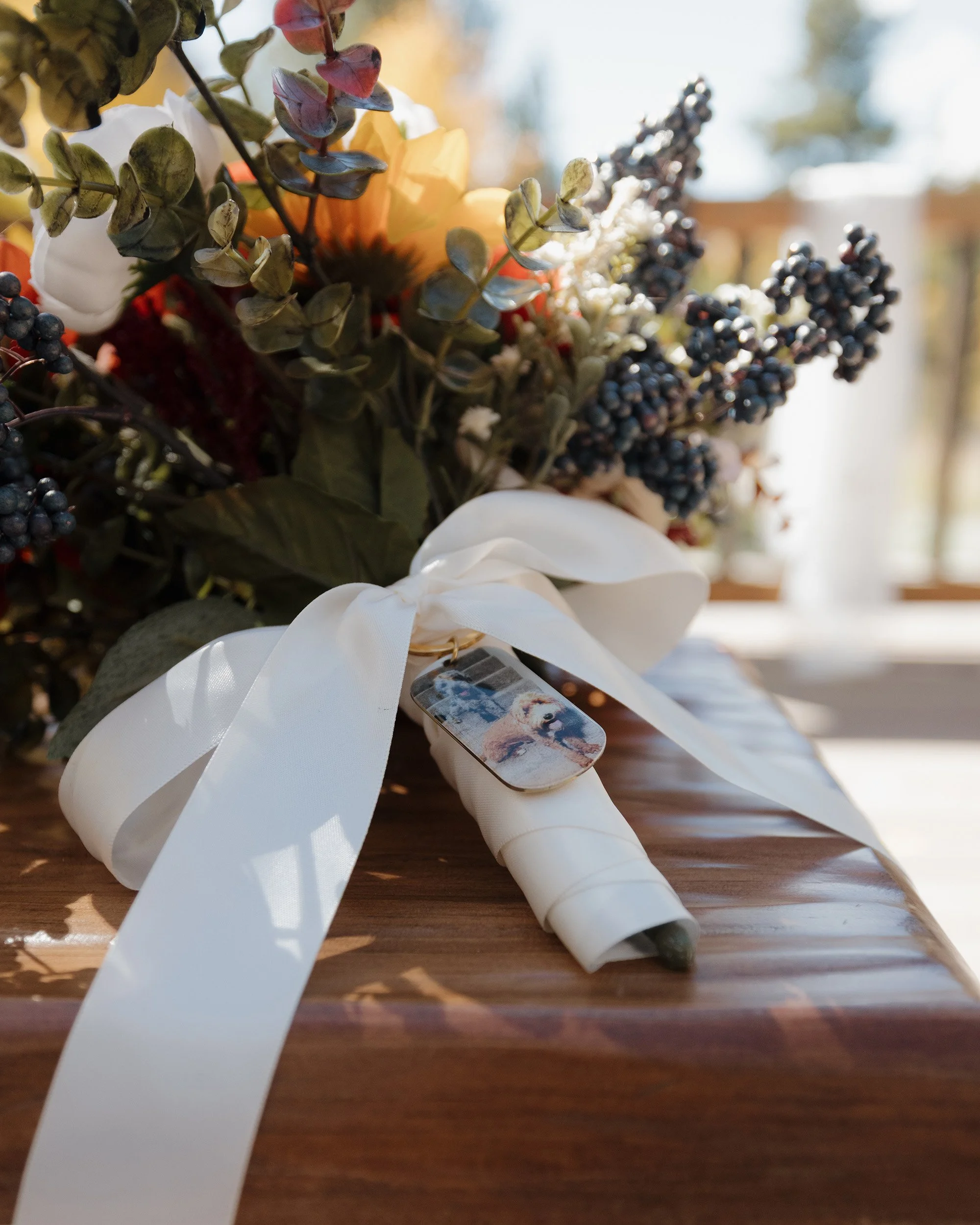 A bouquet of flowers and foliage wrapped with a white ribbon, with a small photo keychain attached, placed on a wooden table with sunlight filtering through a window in the background.