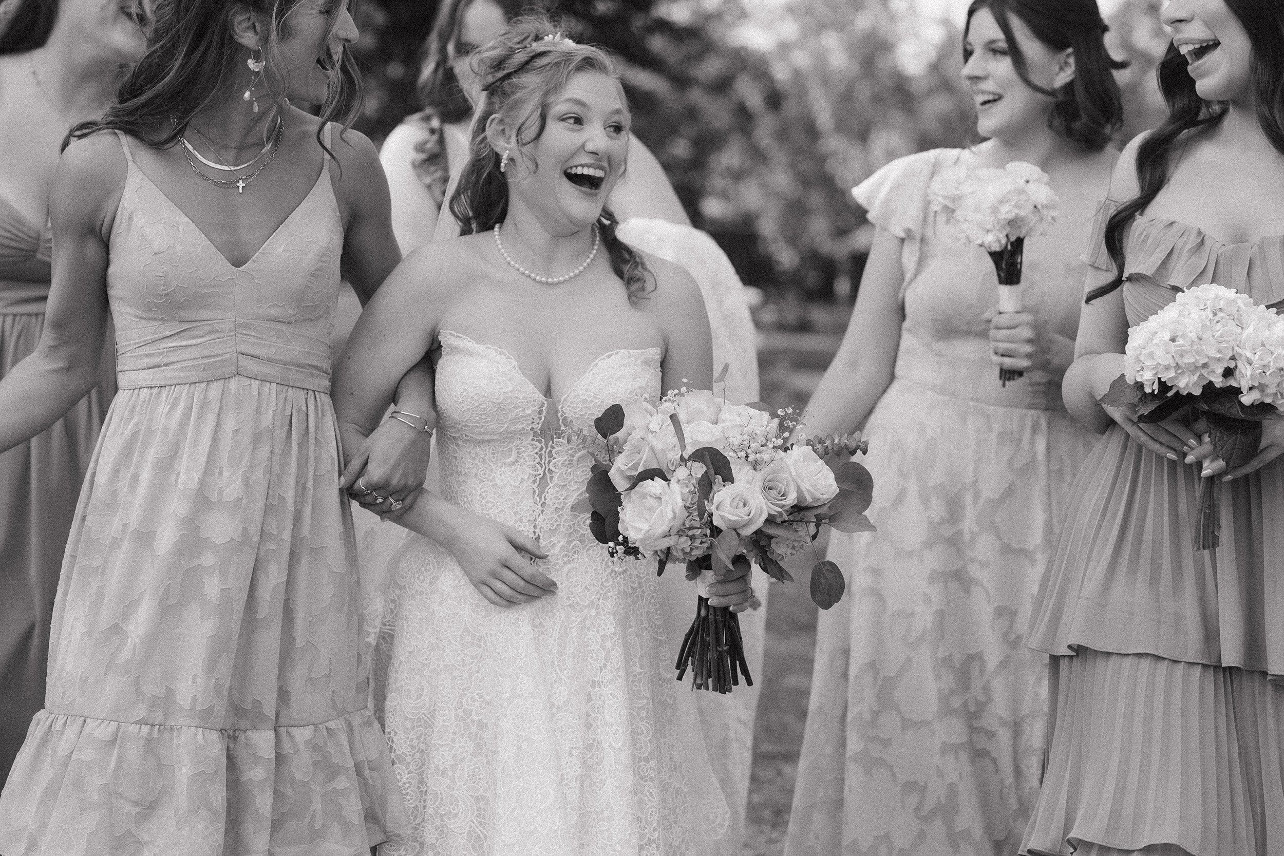 A group of women in dresses celebrating at a wedding, with the bride holding a bouquet of roses and surrounded by bridesmaids holding flowers, smiling and laughing outdoors.