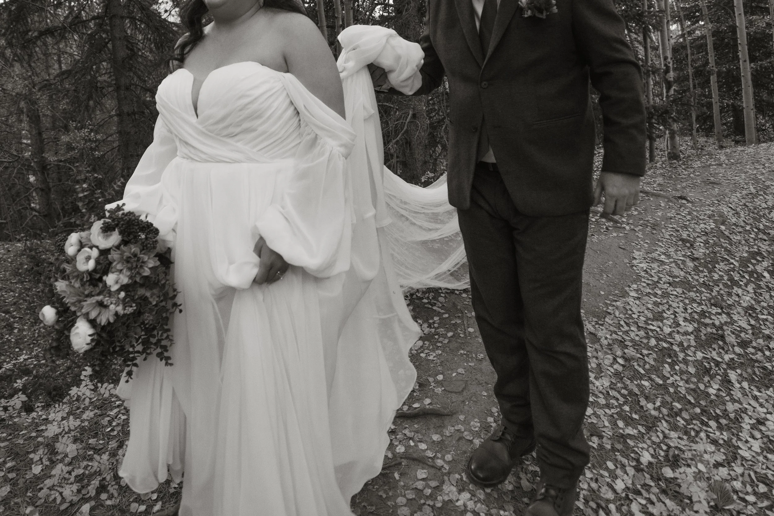A bride in a white wedding dress holding a bouquet of flowers walking in the woods with a groom in a dark suit.