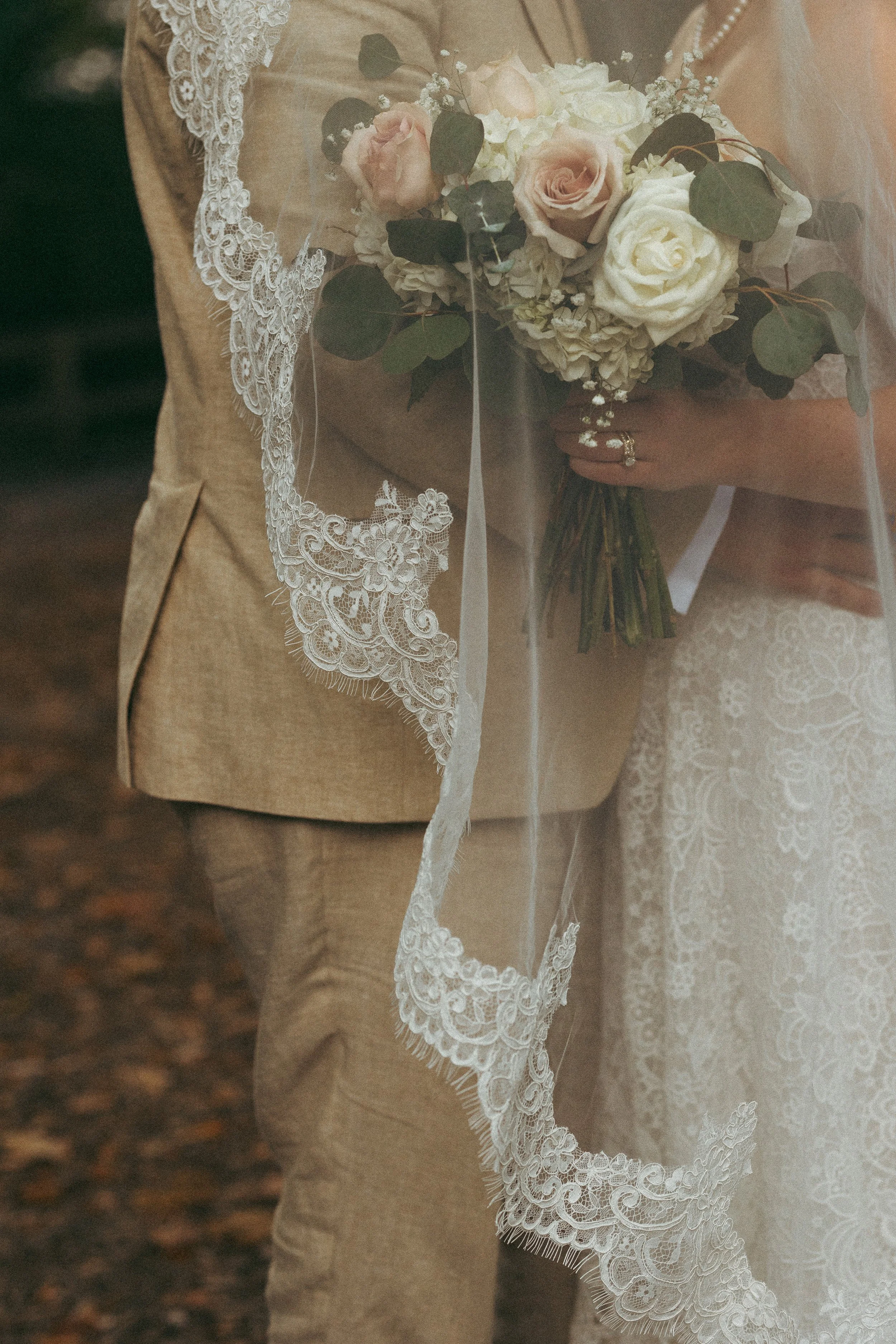 Close-up of a bride holding a bouquet of pink and white roses with greenery, under a lace veil, in front of a person in a beige suit.