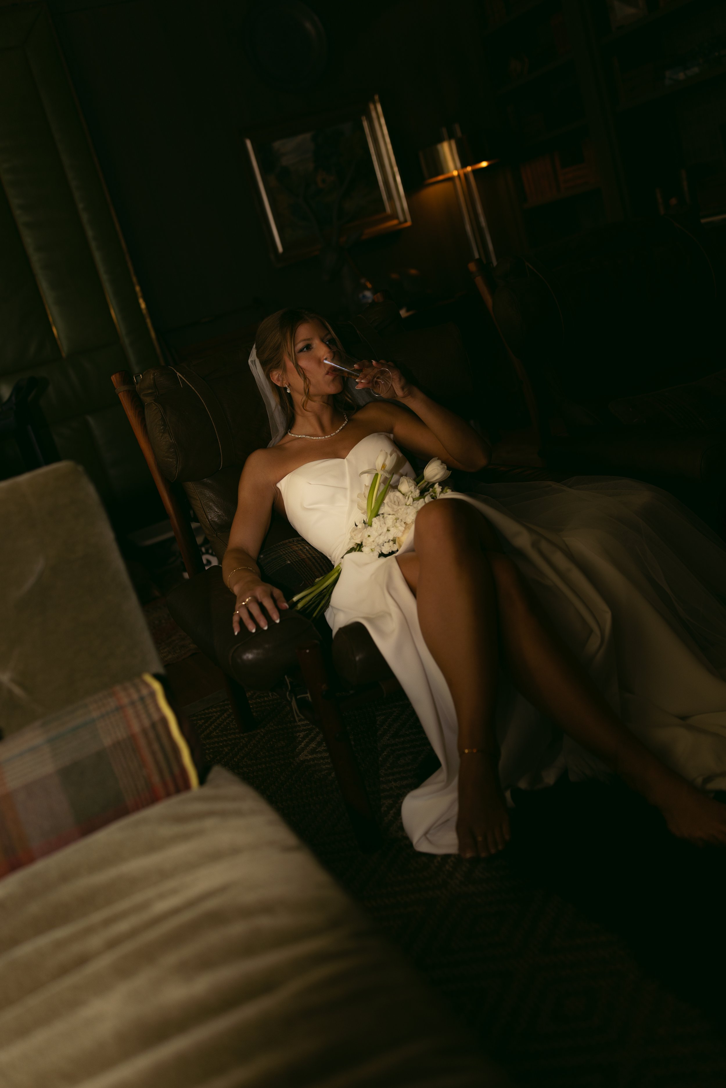 A bride in a strapless white wedding dress lying on a dark leather chair, smoking a cigarette, with a bouquet of white flowers on her lap, in a dimly lit room.