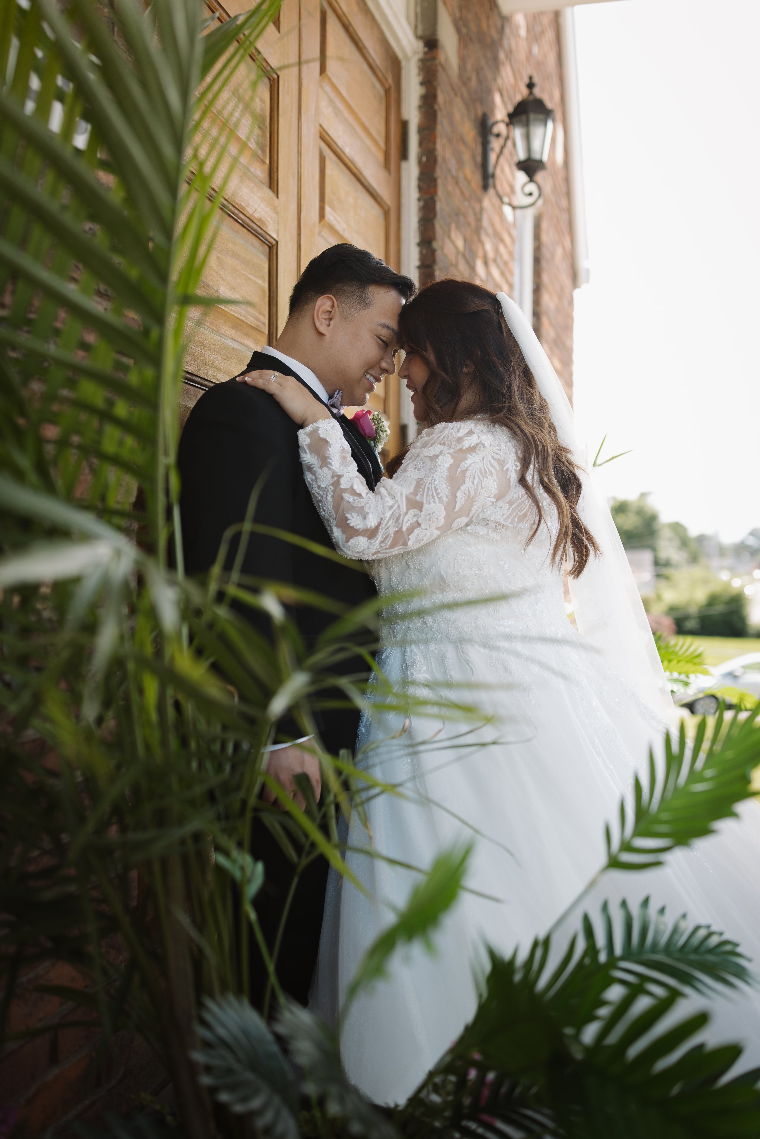 A bride and groom sharing a close, joyful moment outside a brick building, with lush greenery in the foreground.