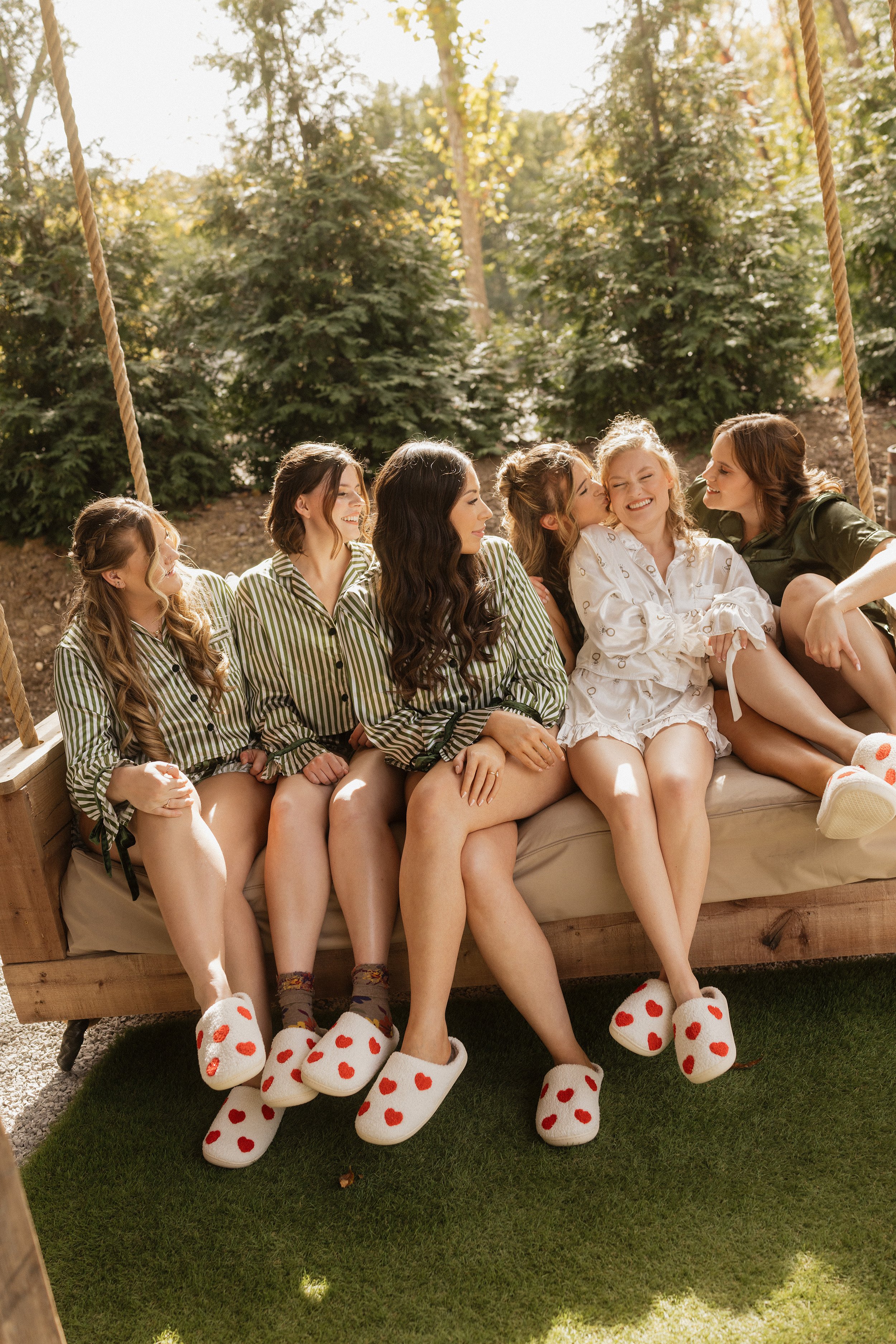 A group of six women sitting on a wooden swing, sharing a moment of laughter and affection outdoors. They are dressed casually, with some wearing matching striped pajamas and slippers with red hearts.