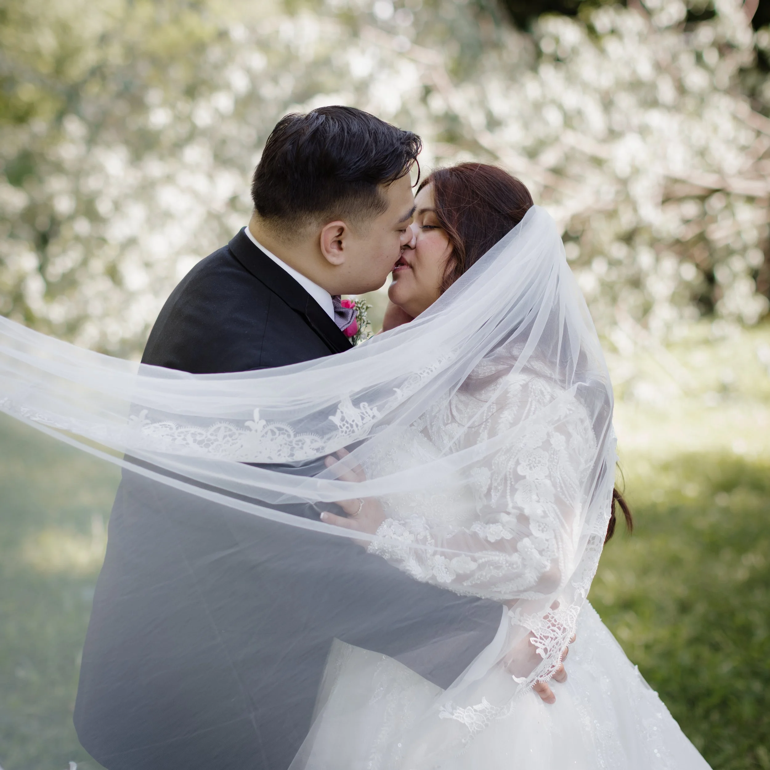 A bride and groom sharing a kiss outdoors, with the bride wearing a lace wedding dress and veil, and the groom in a black tuxedo, under a softly blurred background of trees.