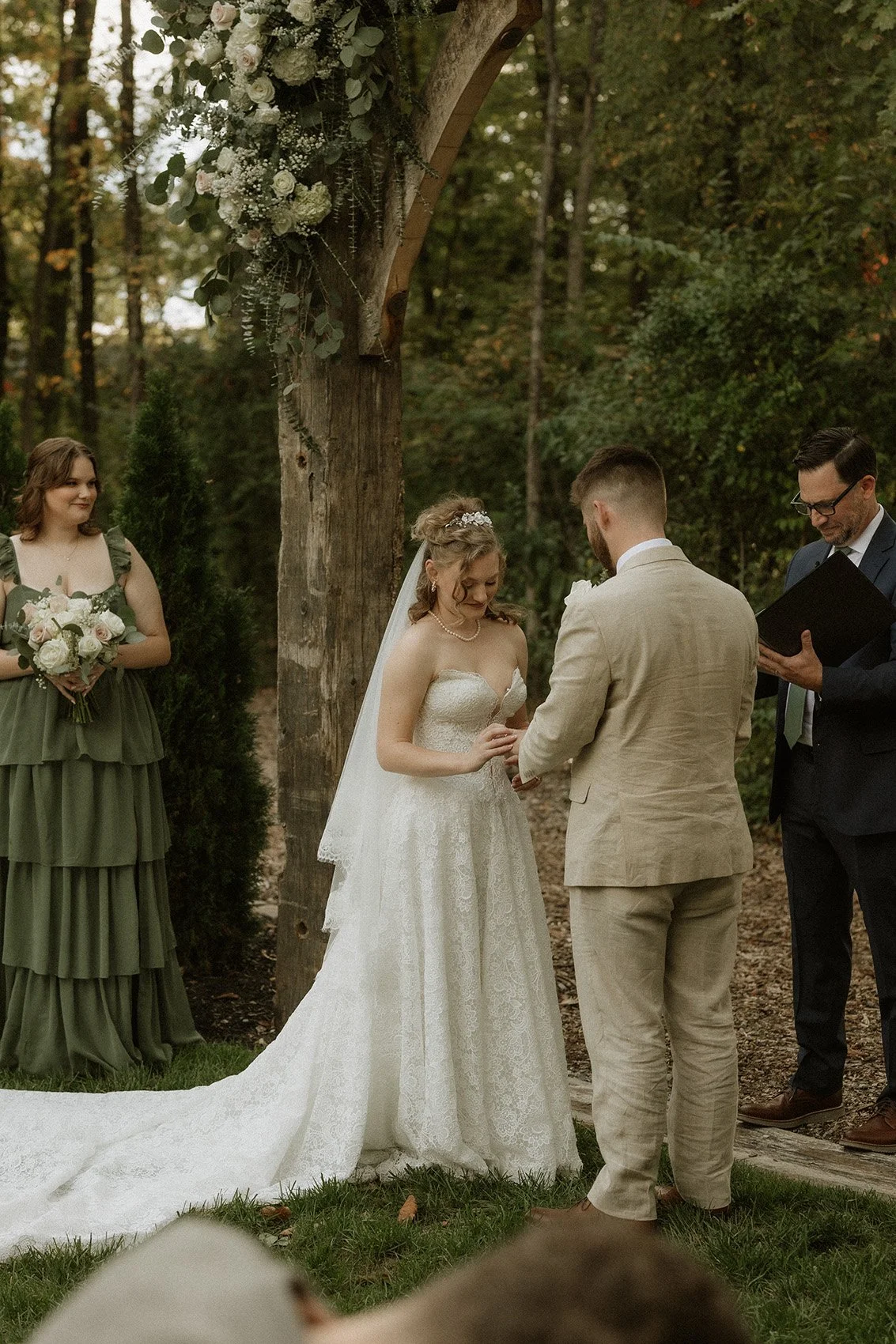 A couple exchanging vows at an outdoor wedding ceremony under a wooden arch decorated with flowers, with a bridesmaid holding a bouquet standing nearby.