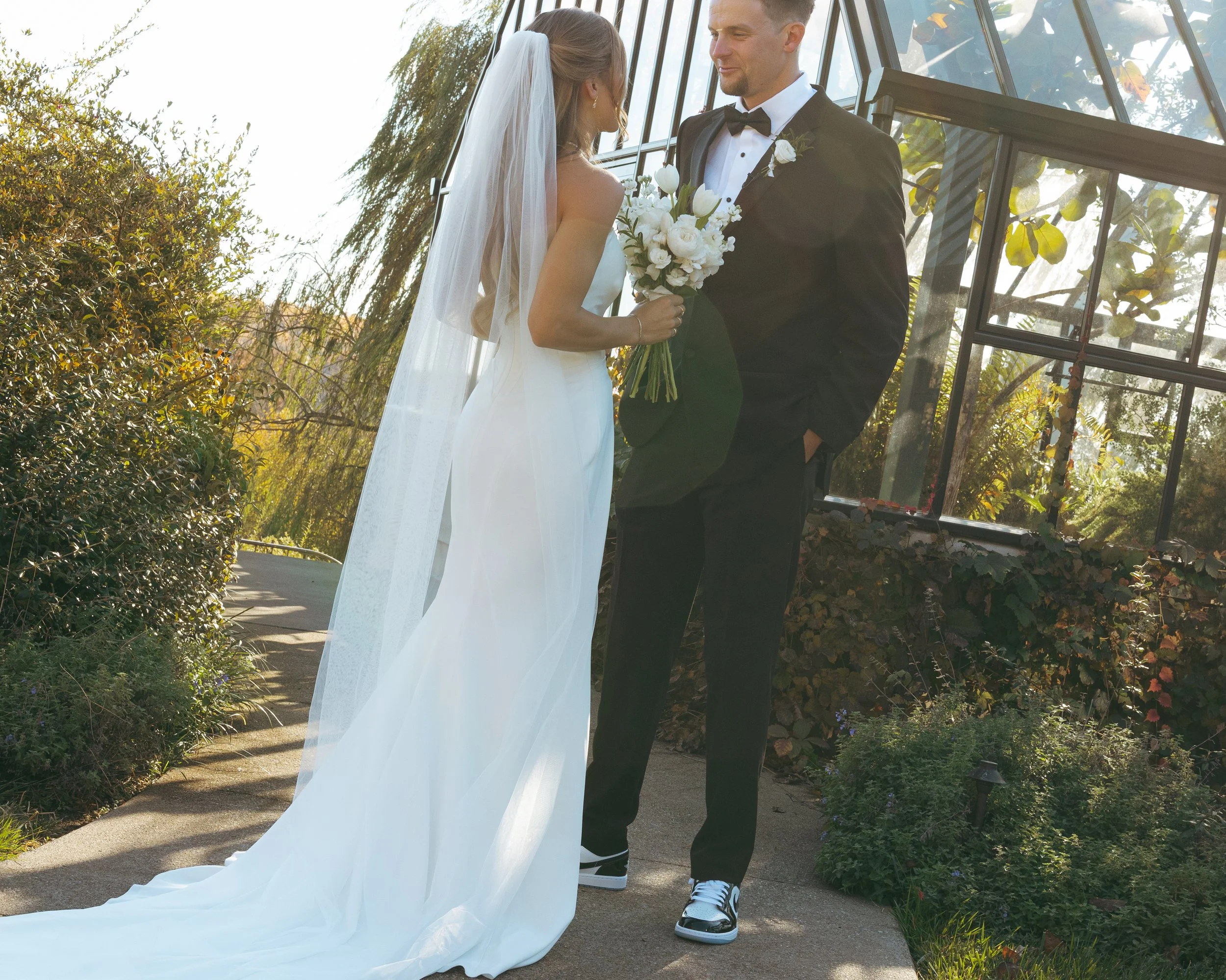 A bride and groom standing outdoors, facing each other, with the bride holding a bouquet of white flowers. The bride is wearing a white wedding gown and veil, while the groom is dressed in a black tuxedo with a bow tie. They are smiling at each other