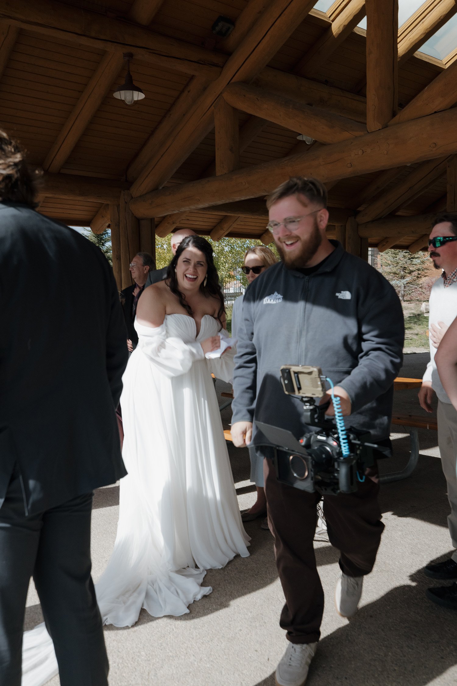 A group of people smiling and laughing at what appears to be a wedding celebration, with a woman in a white wedding dress and a man in casual clothing under a wooden pavilion.