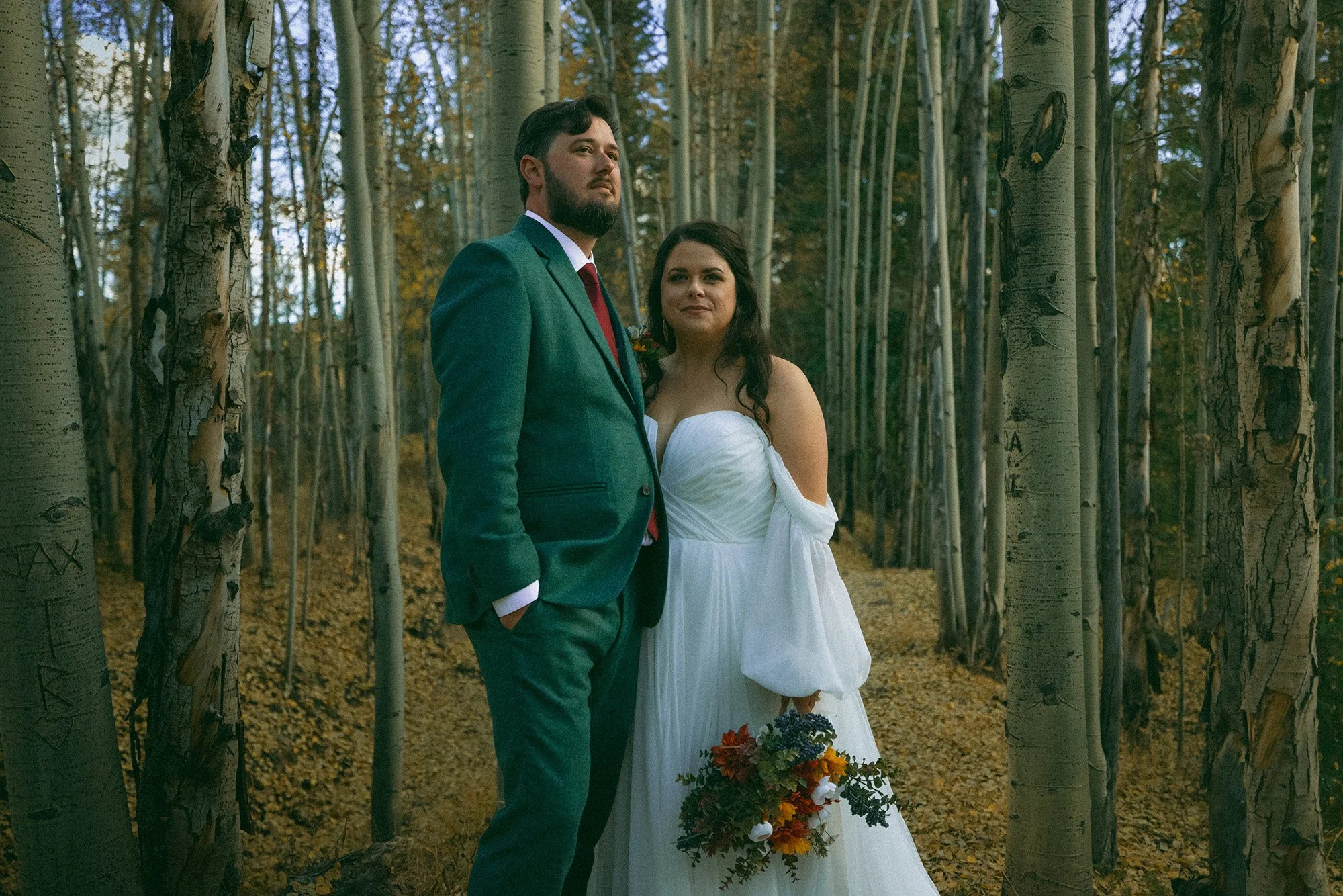 A man and woman in wedding attire standing in a forest with tall trees and autumn leaves.