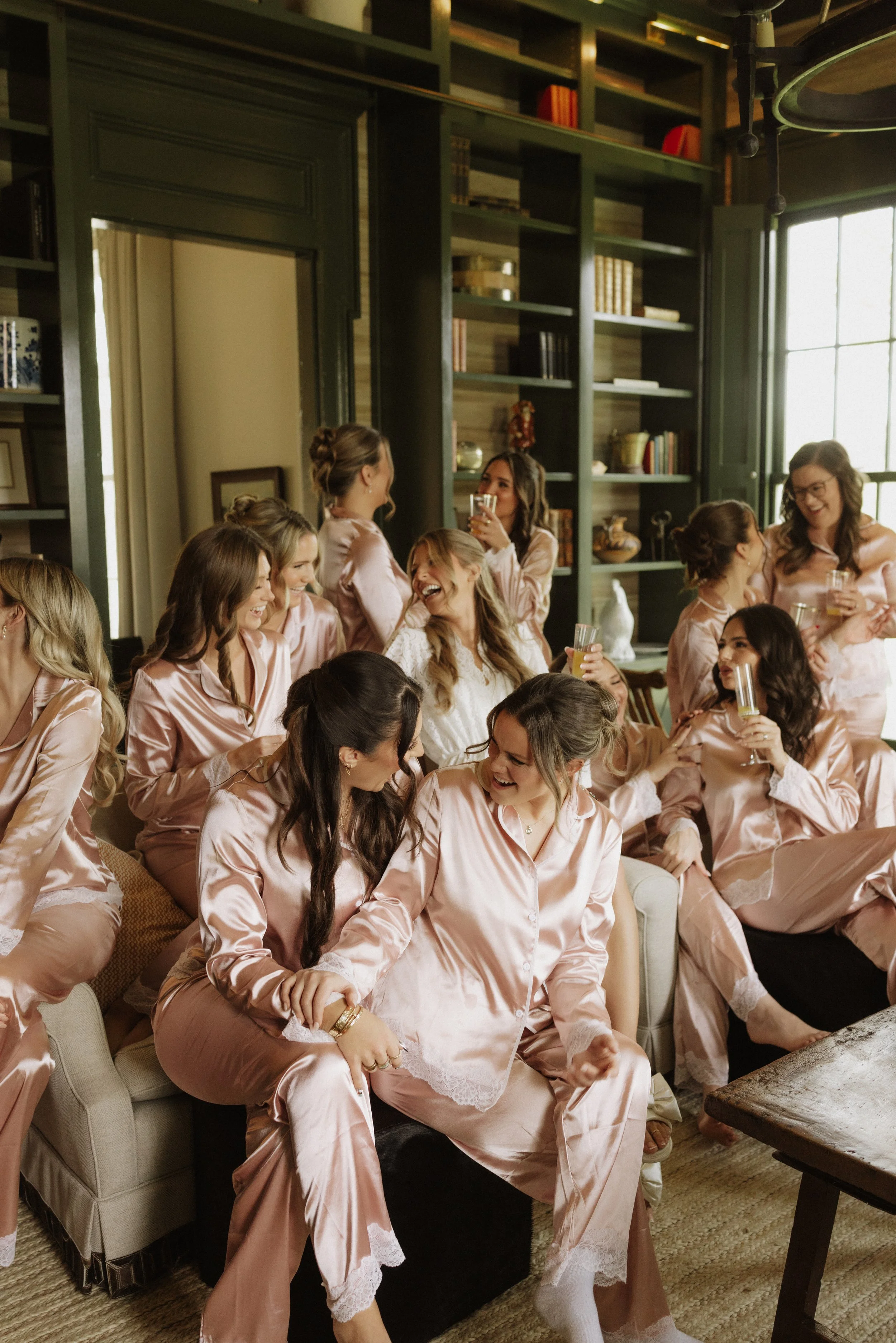 A group of women wearing matching pink satin pajamas are sitting and standing in a cozy, well-lit living room, celebrating, laughing, and holding drinks.