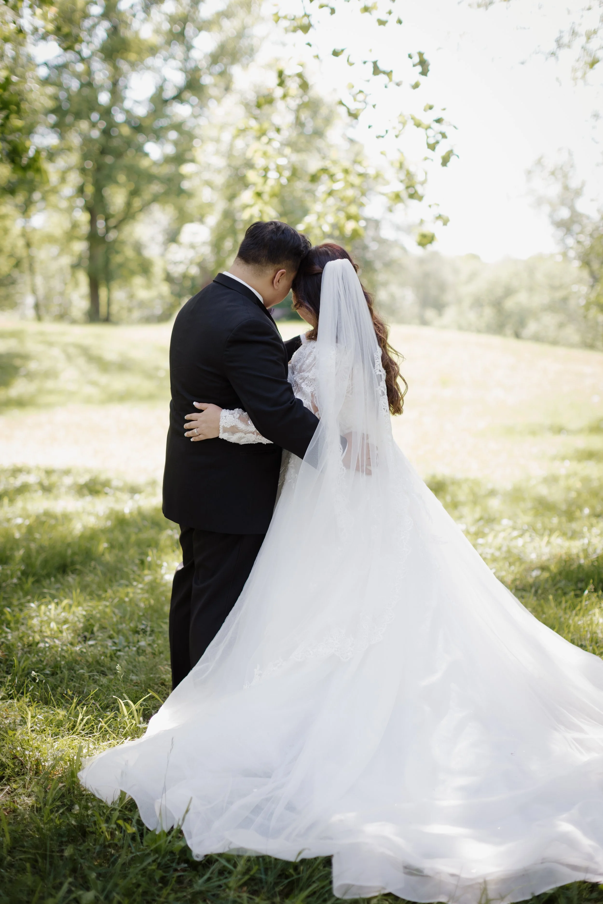 A bride and groom standing close together in a grassy park, embracing with their foreheads touching, surrounded by trees on a sunny day.