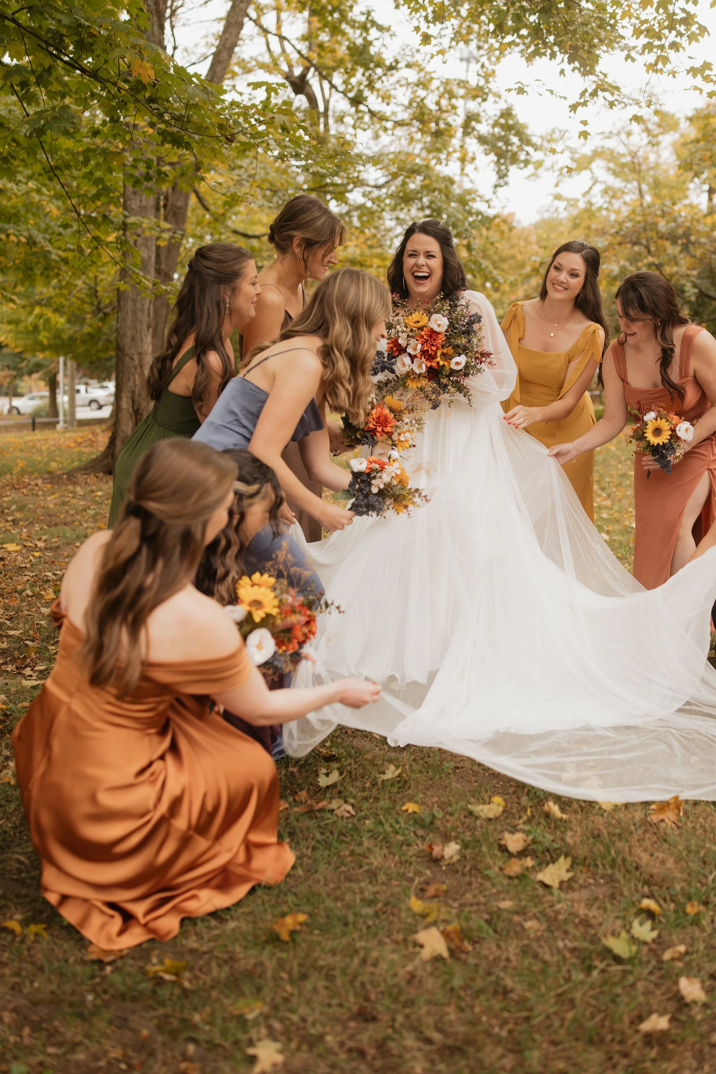 A woman wearing a white wedding dress is surrounded by several bridesmaids wearing colorful dresses, holding bouquets, as they celebrate outdoors during fall with autumn leaves and trees in the background.