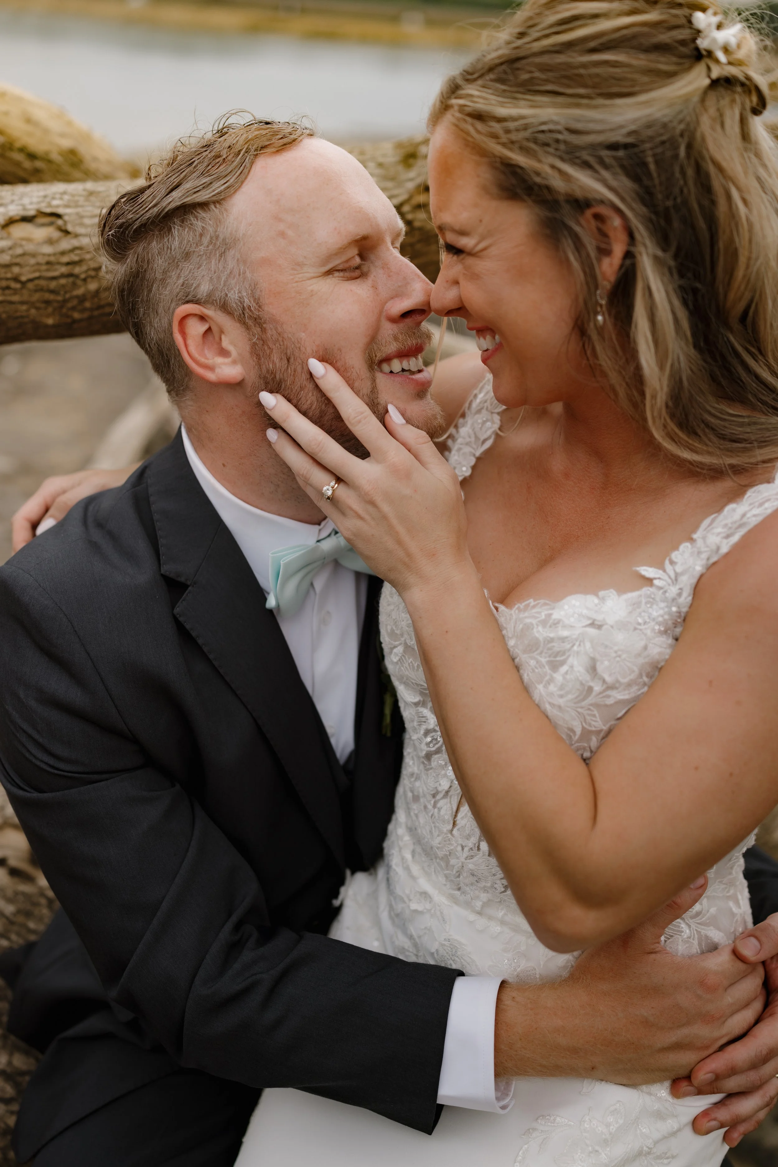 A bride and groom in wedding attire share an intimate moment outdoors, with their faces close together, smiling, and the bride gently touching the groom's face, while the groom holds the bride's waist.