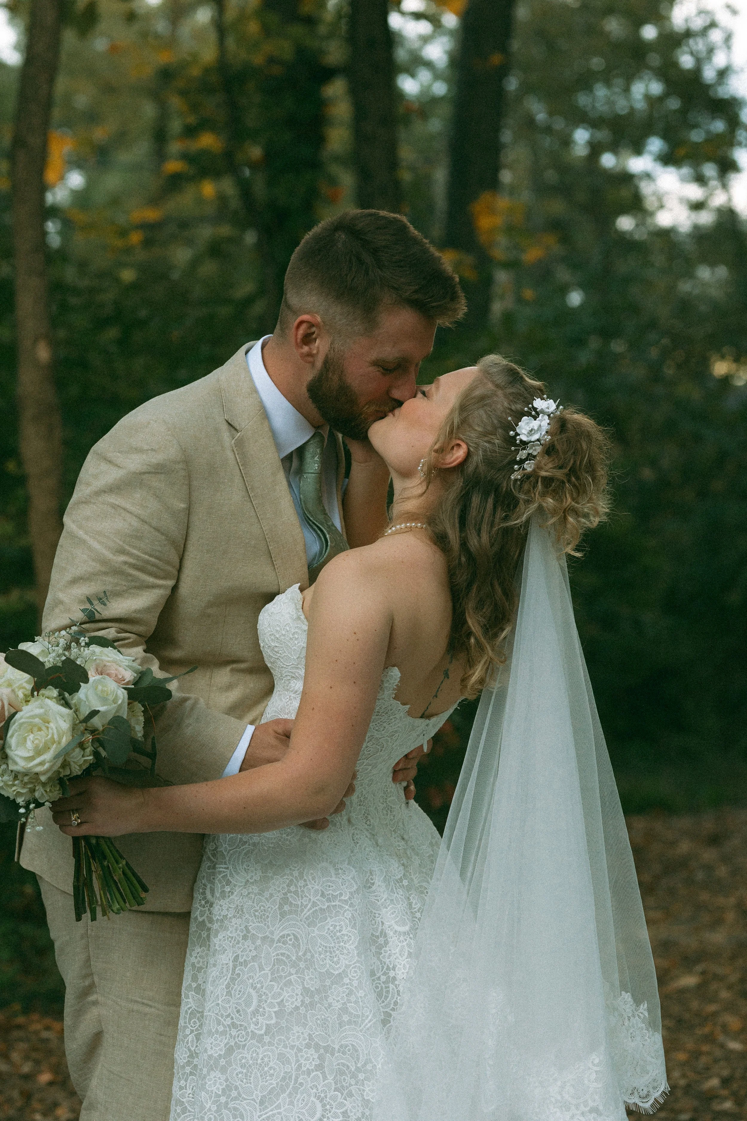 A newlywed couple sharing a kiss outdoors in a forest setting during their wedding.