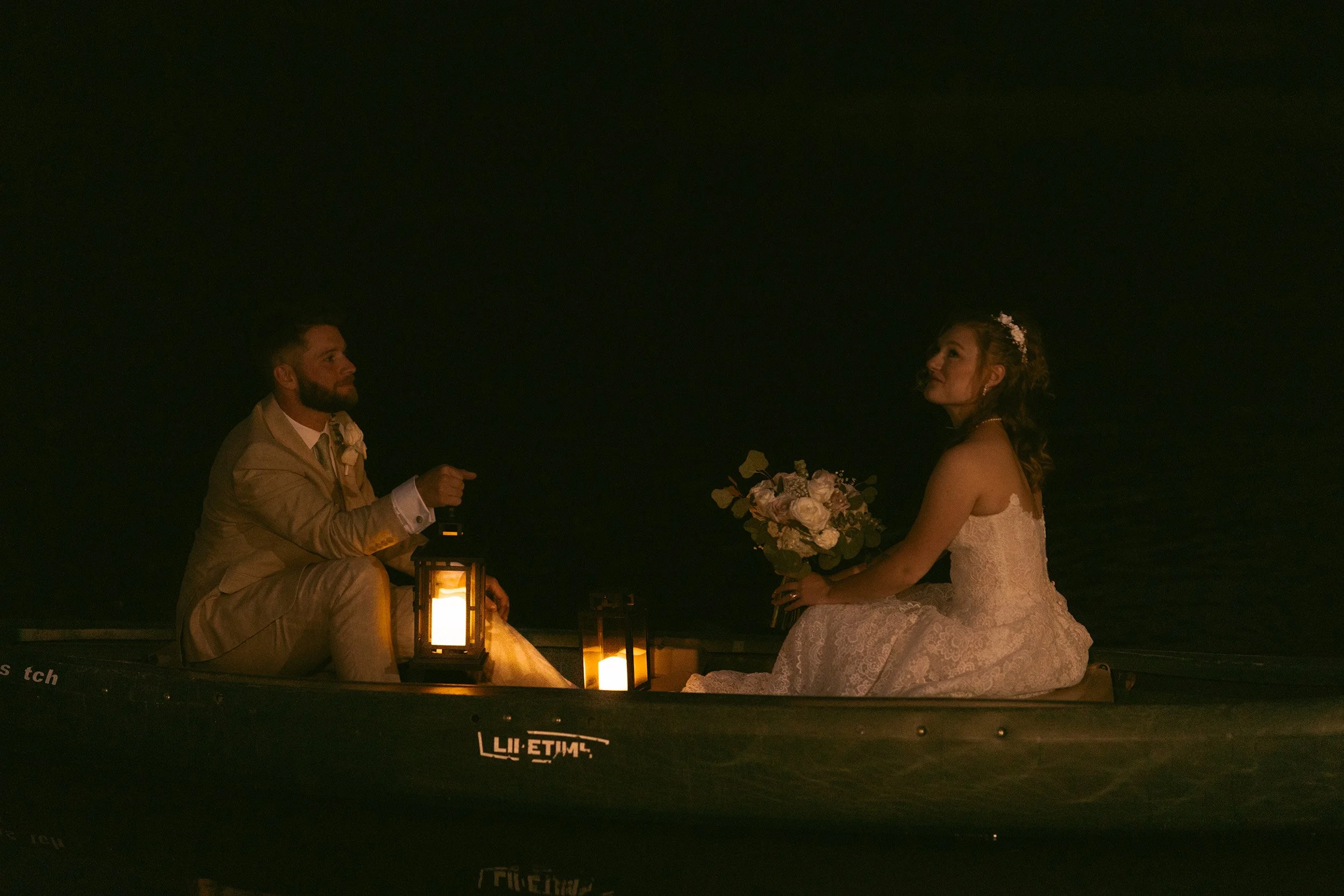 A bride and groom sitting in a boat at night, illuminated by lanterns, with the bride holding a bouquet of flowers, enjoying a romantic moment.
