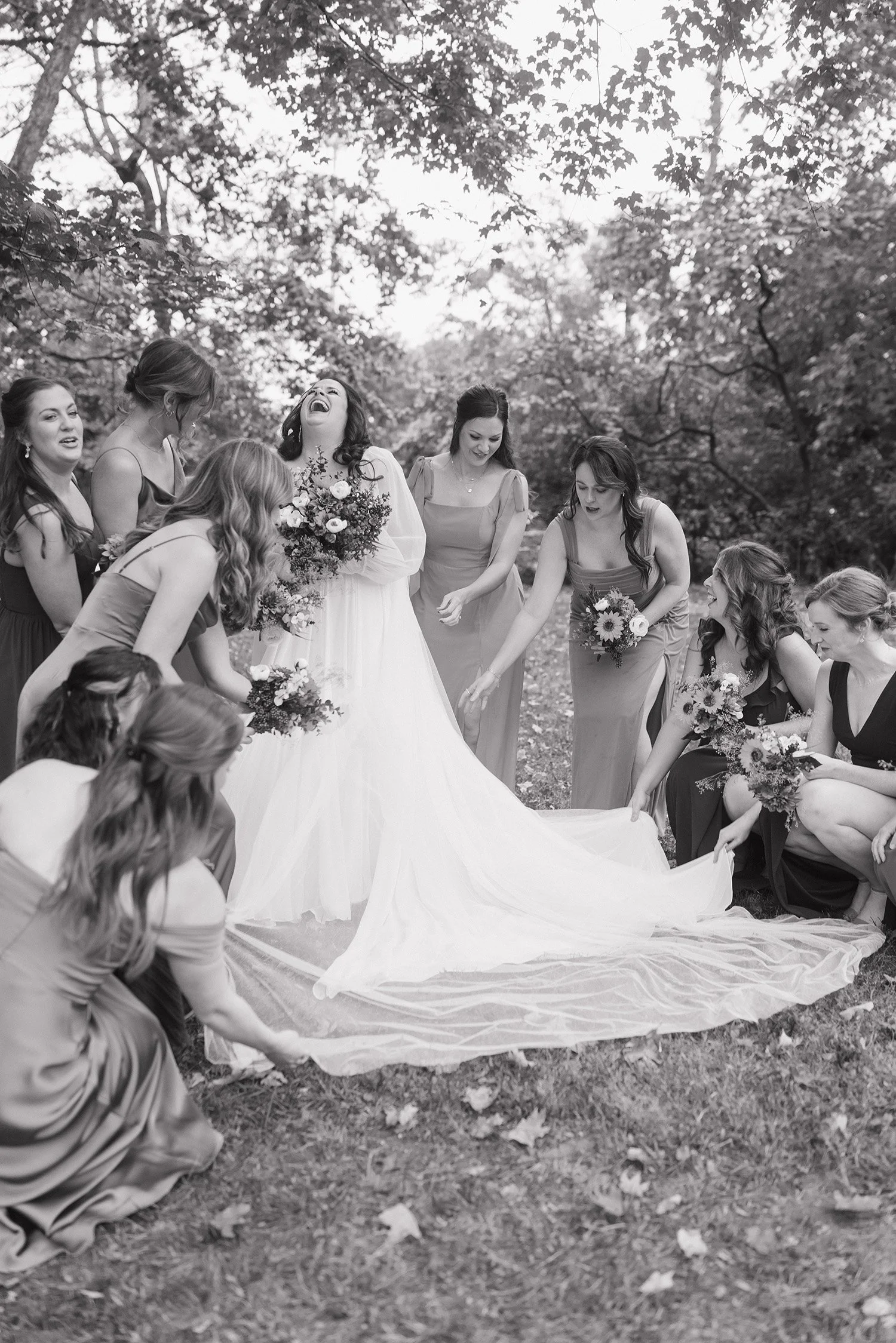 Group of women in dresses, with one bride in a white dress holding a bouquet, surrounded by bridesmaids outdoors, with trees and leaves in the background.