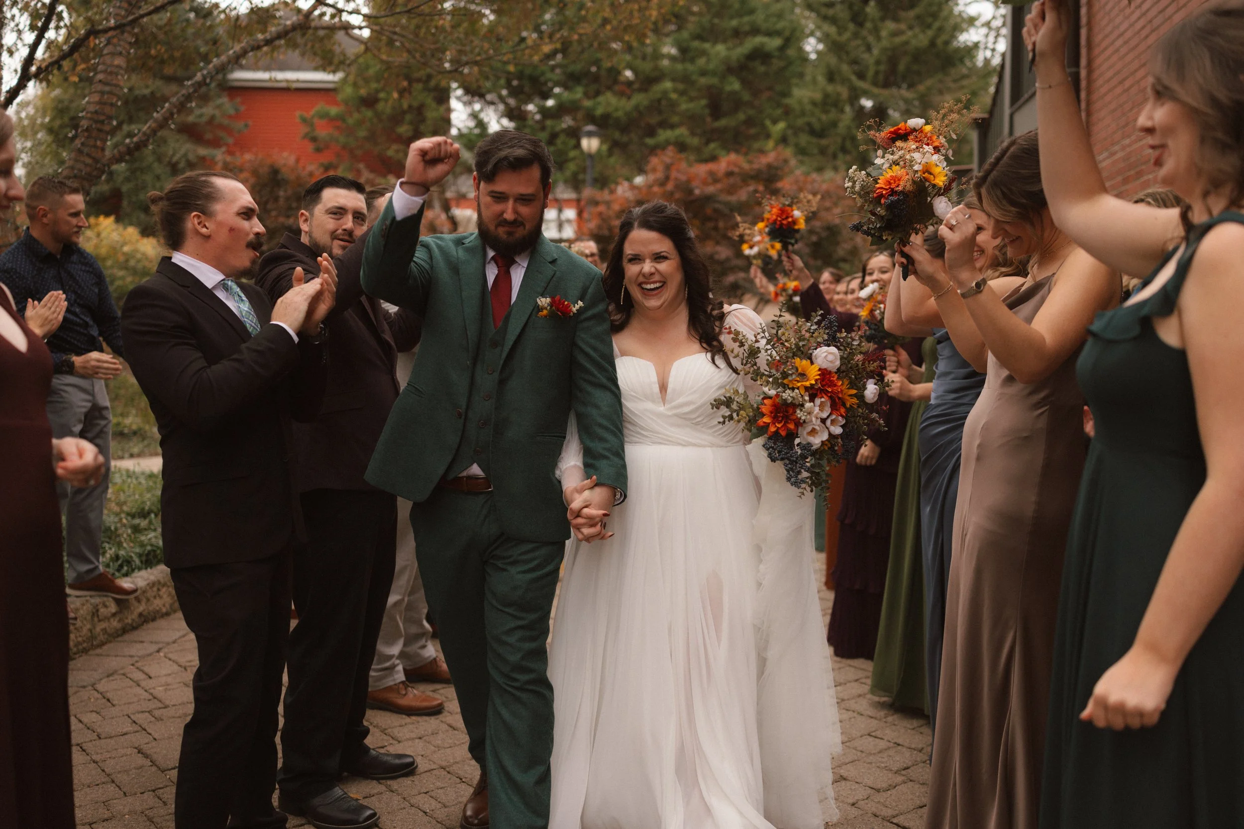 A bride and groom holding hands, walking through a crowd of people celebrating outdoors with fall foliage in the background.