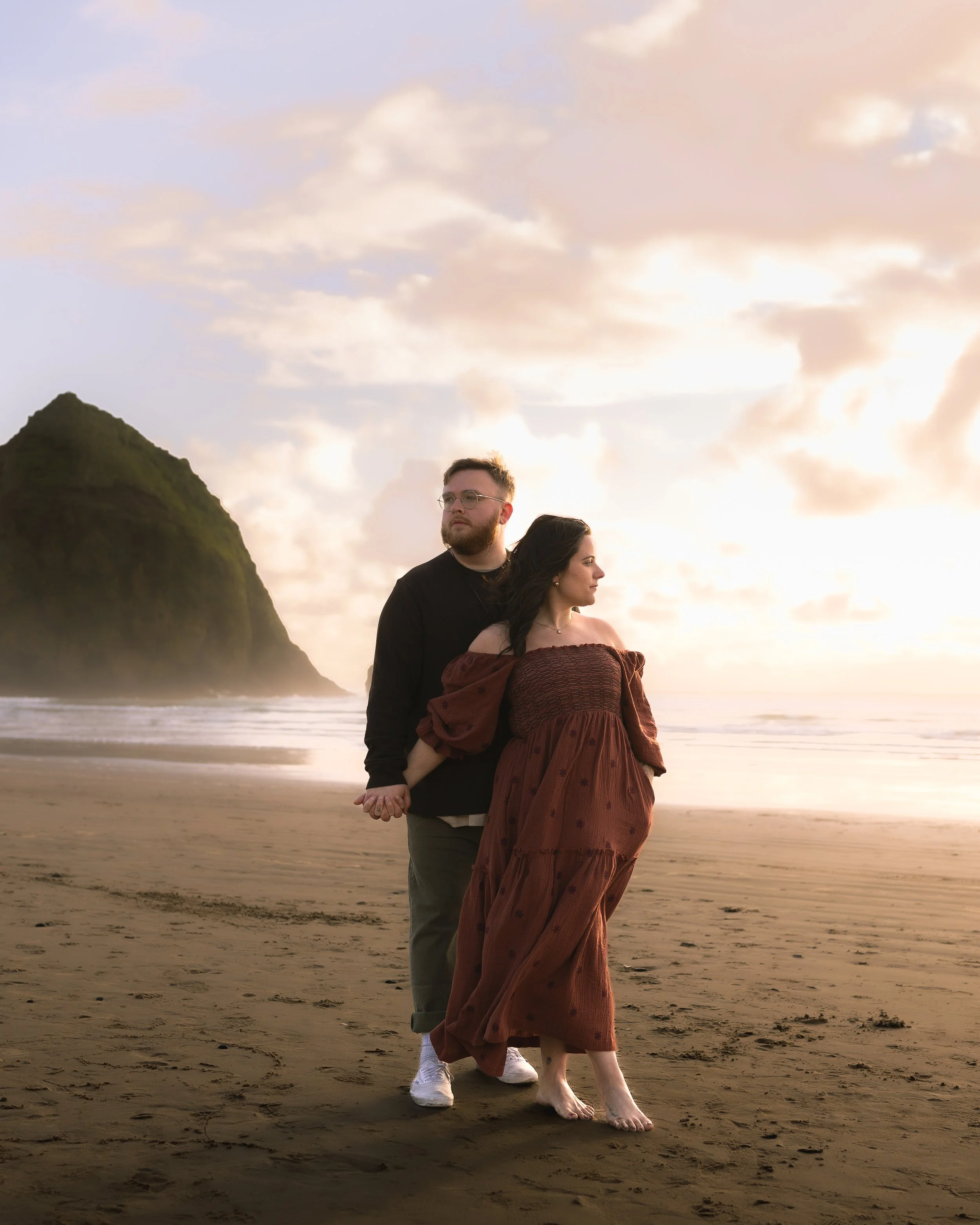 A couple holding hands on the beach during sunset, with the woman wearing an off-shoulder brown dress and the man in dark clothing, a large green cliff in the background, and a partly cloudy sky.