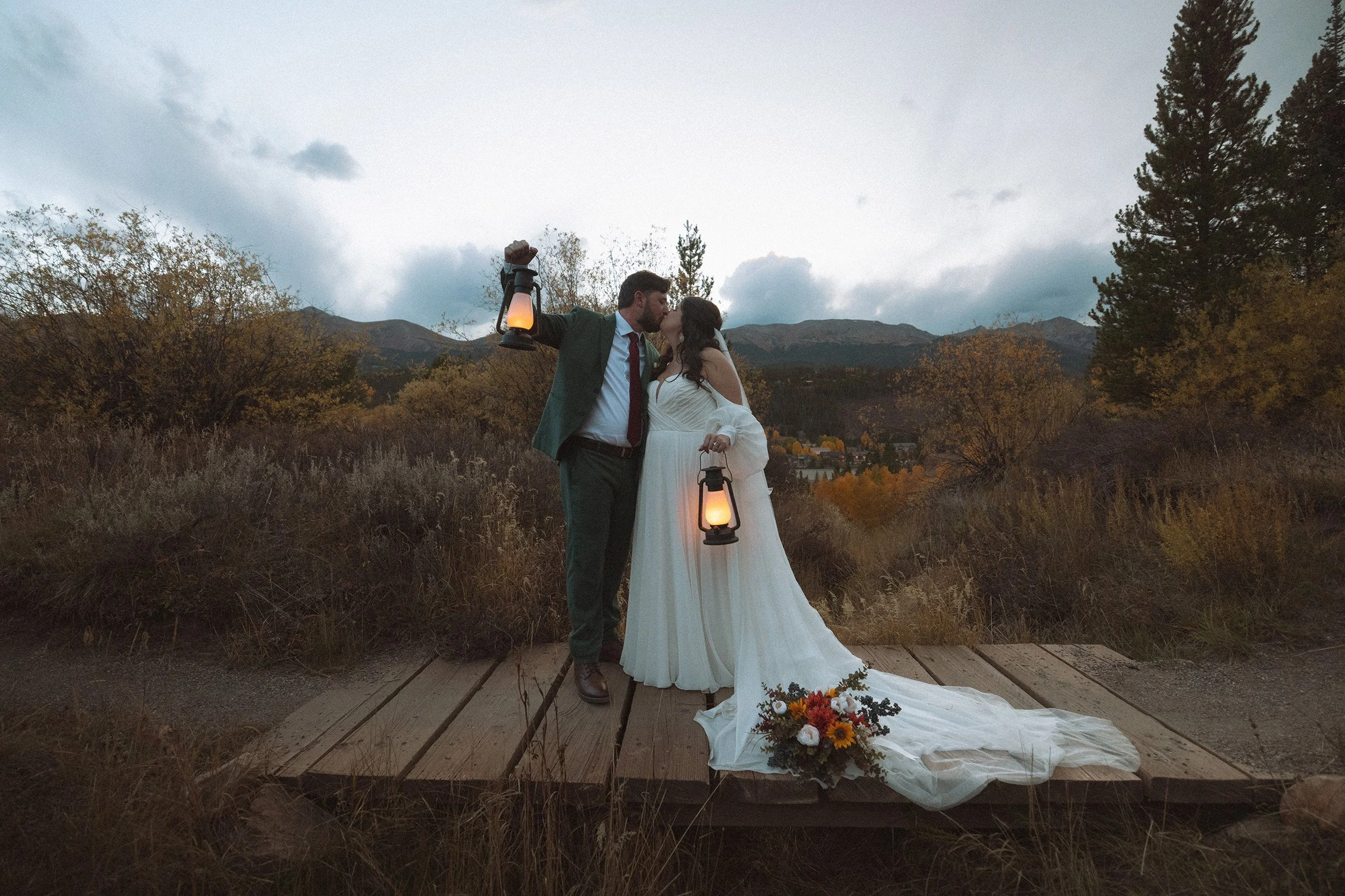 A couple in wedding attire kissing on a wooden platform outdoors, holding lanterns, with a bouquet of flowers nearby, surrounded by trees and mountains.