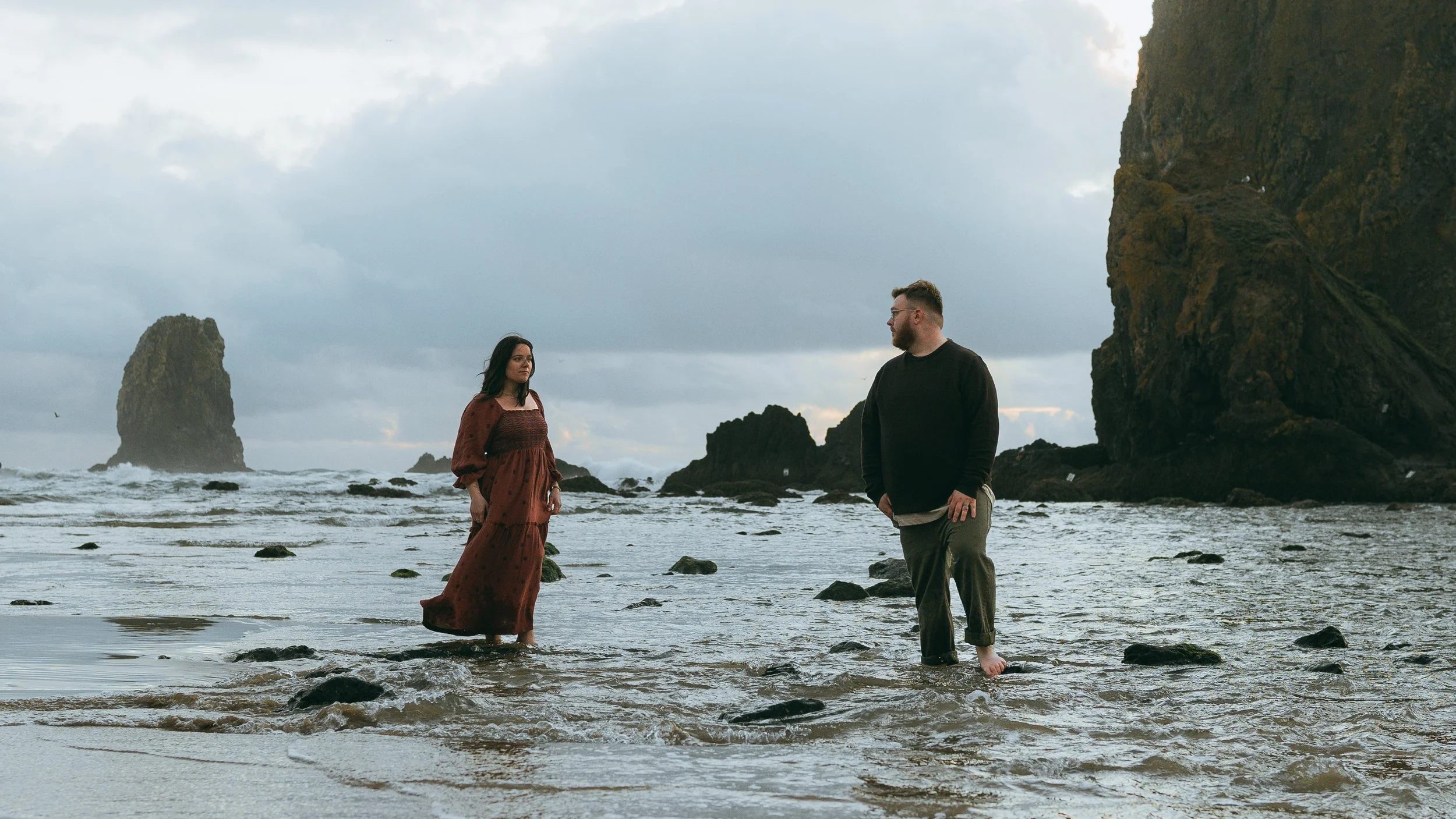 A woman in a long brown dress and a man in a black sweater and gray pants standing in shallow ocean water near rocky formations with a cloudy sky overhead.