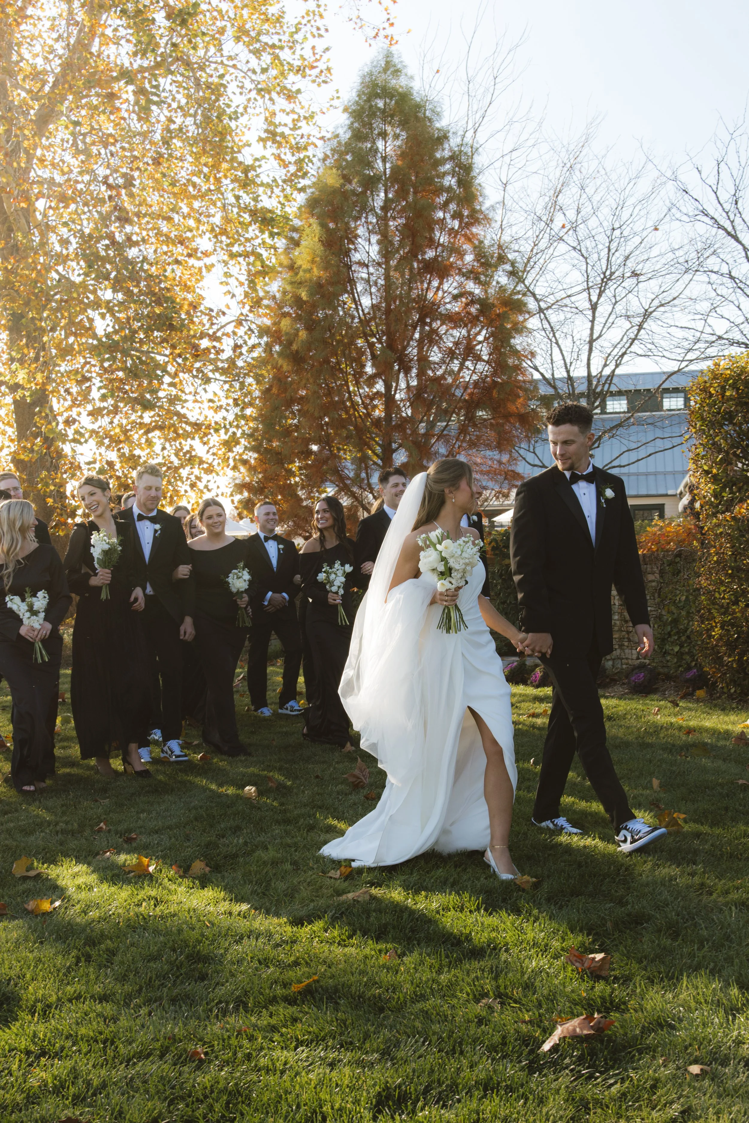 Wedding party outdoors with bride and groom walking on grass, guests in black attire behind them, autumn trees, and sunlight in the background.
