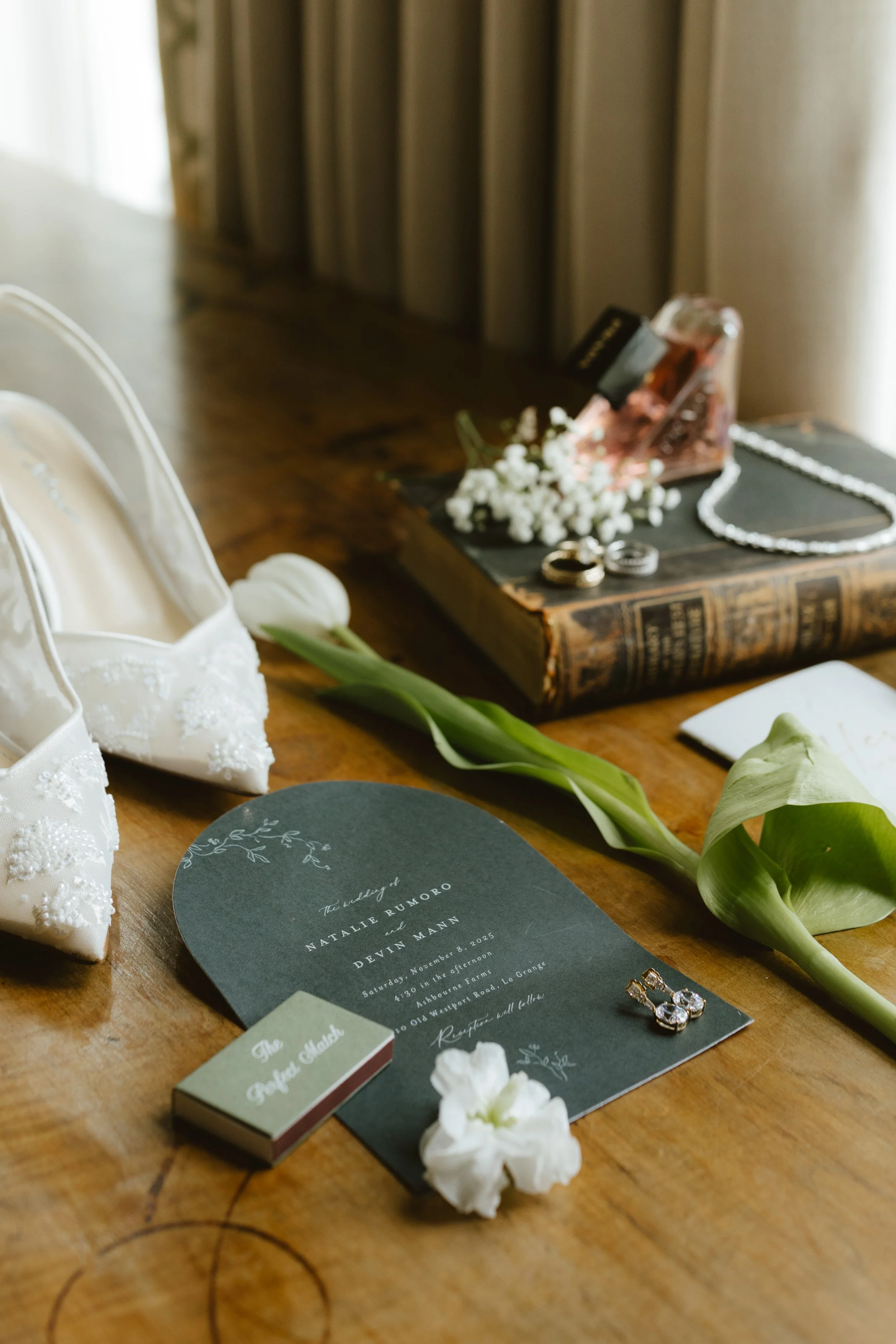 Wedding invitation on a wooden table surrounded by white flowers, jewelry, a pair of white embroidered shoes, a book, and a camera.