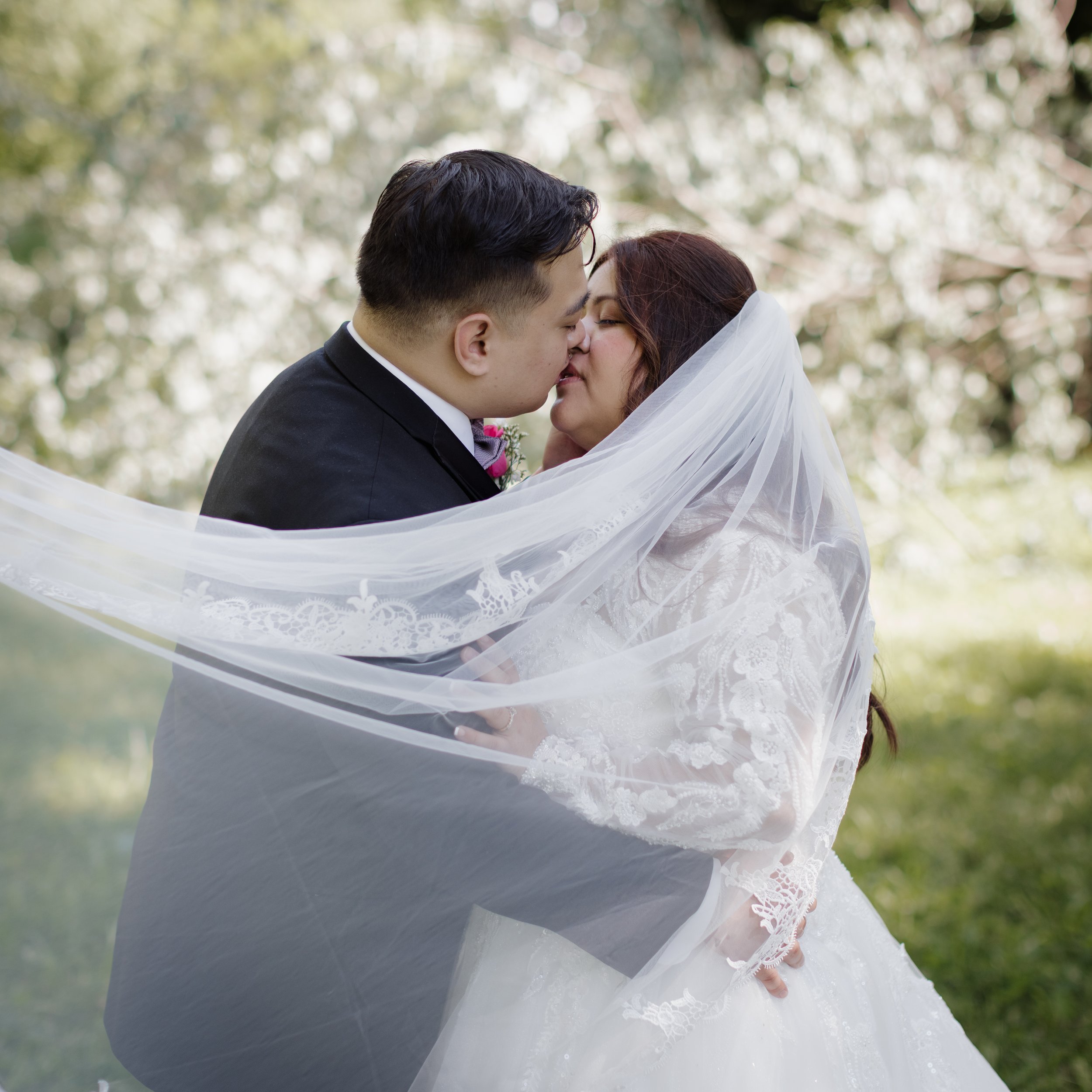 A bride and groom kiss outdoors, with the bride wearing a white lace wedding dress and veil, and the groom in a black tuxedo with a pink boutonniere, surrounded by trees.