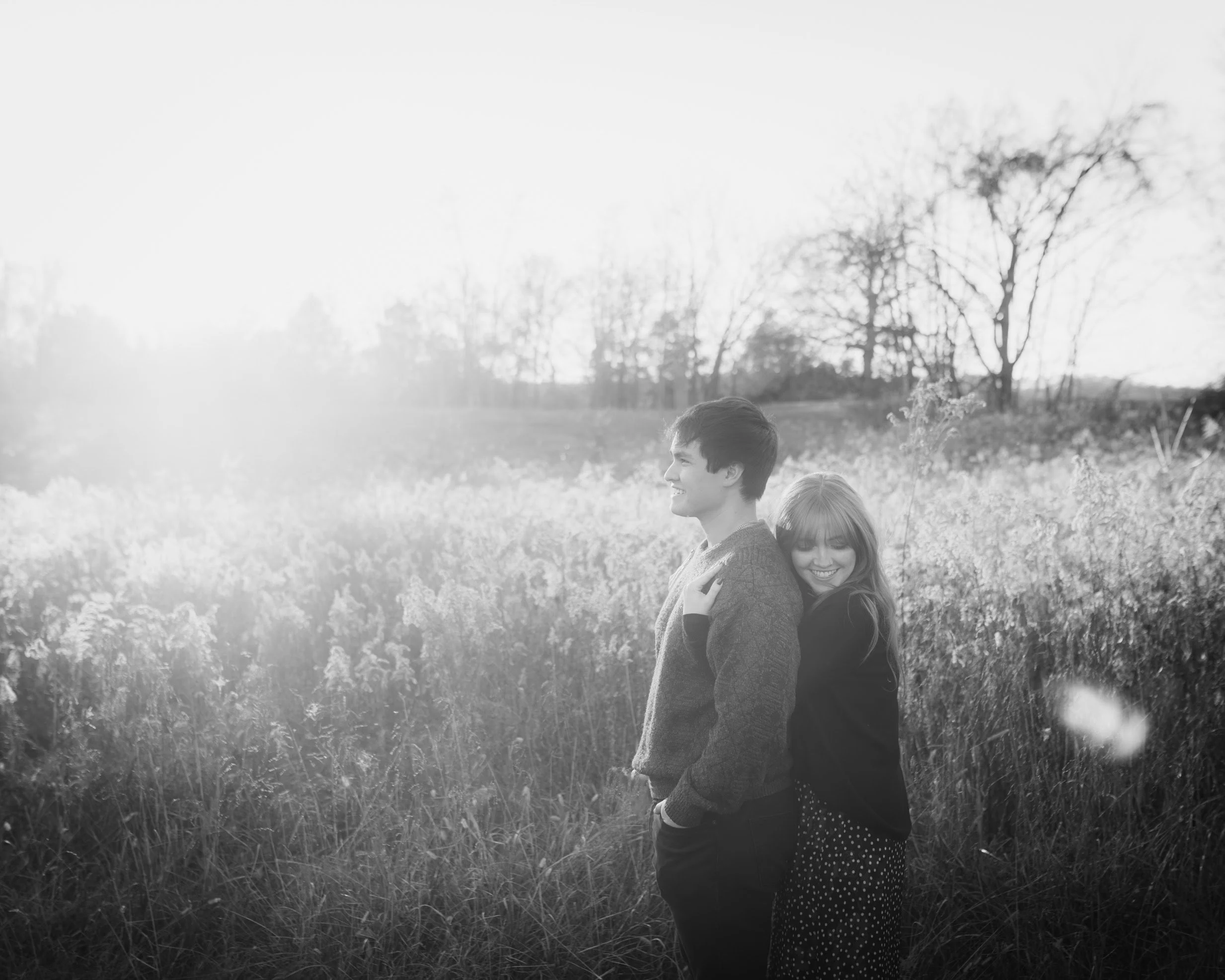 A black and white photo of a young man and woman standing in a field of tall grass, back to back, smiling. The woman is hugging the man from behind, and both are enjoying the outdoor scenery with trees in the background.