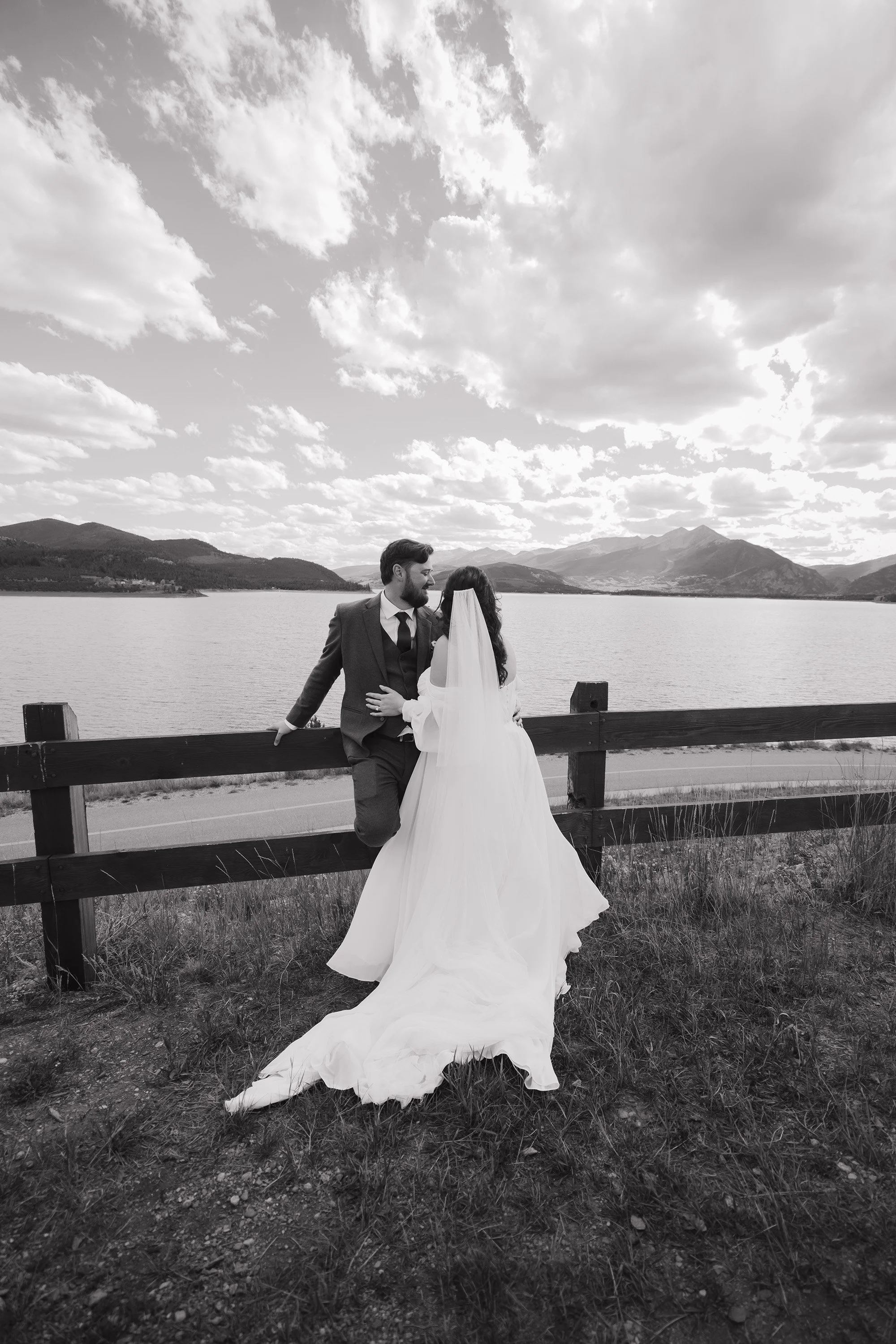 A black and white photo of a bride and groom standing by a wooden fence by a lake, with mountains in the background. The groom, wearing a suit, leans on the fence, and the bride, in a wedding dress, faces him, with her hand on his chest, sharing a ki
