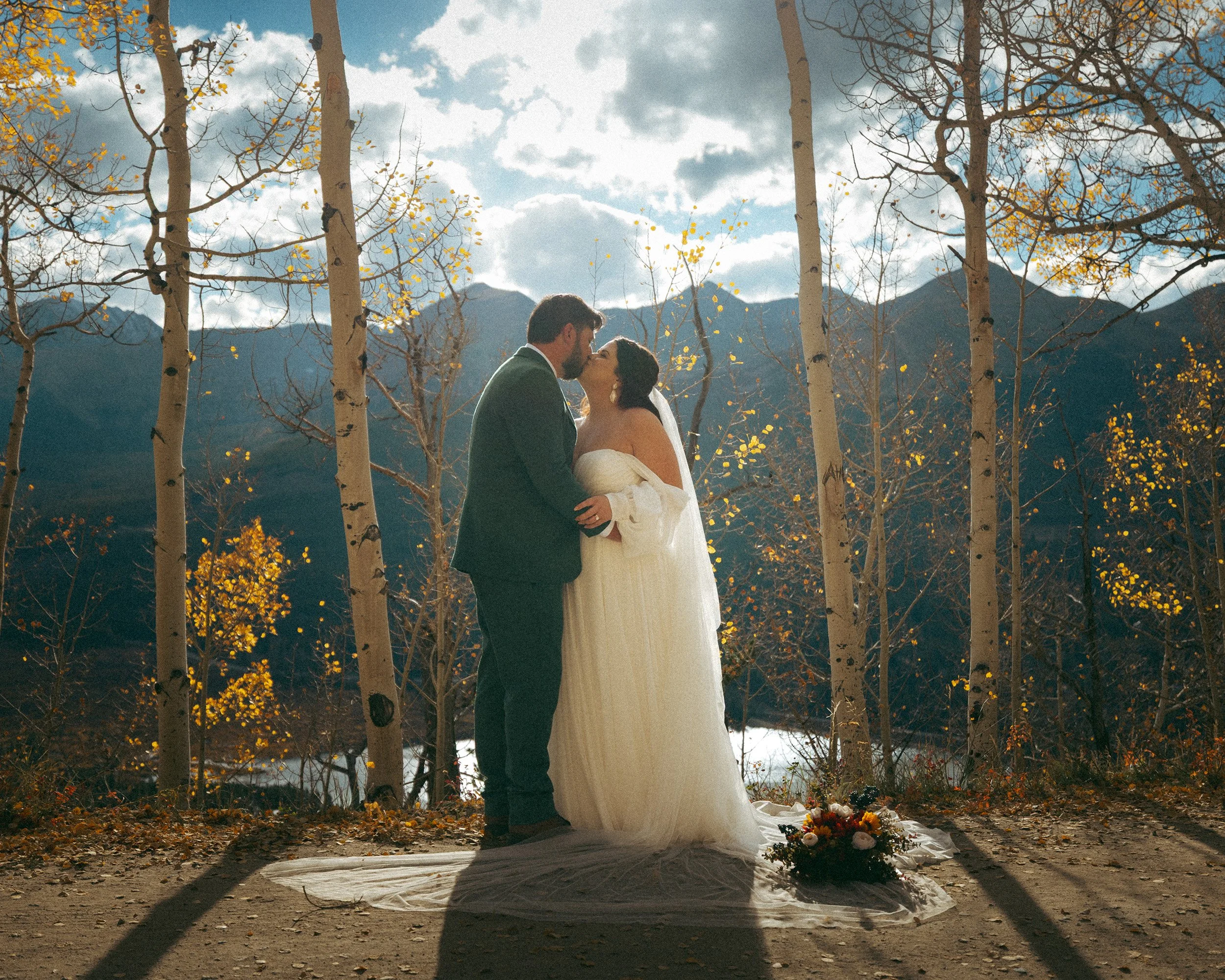 A couple in wedding attire sharing a kiss outdoors among leafless trees with mountains in the background.