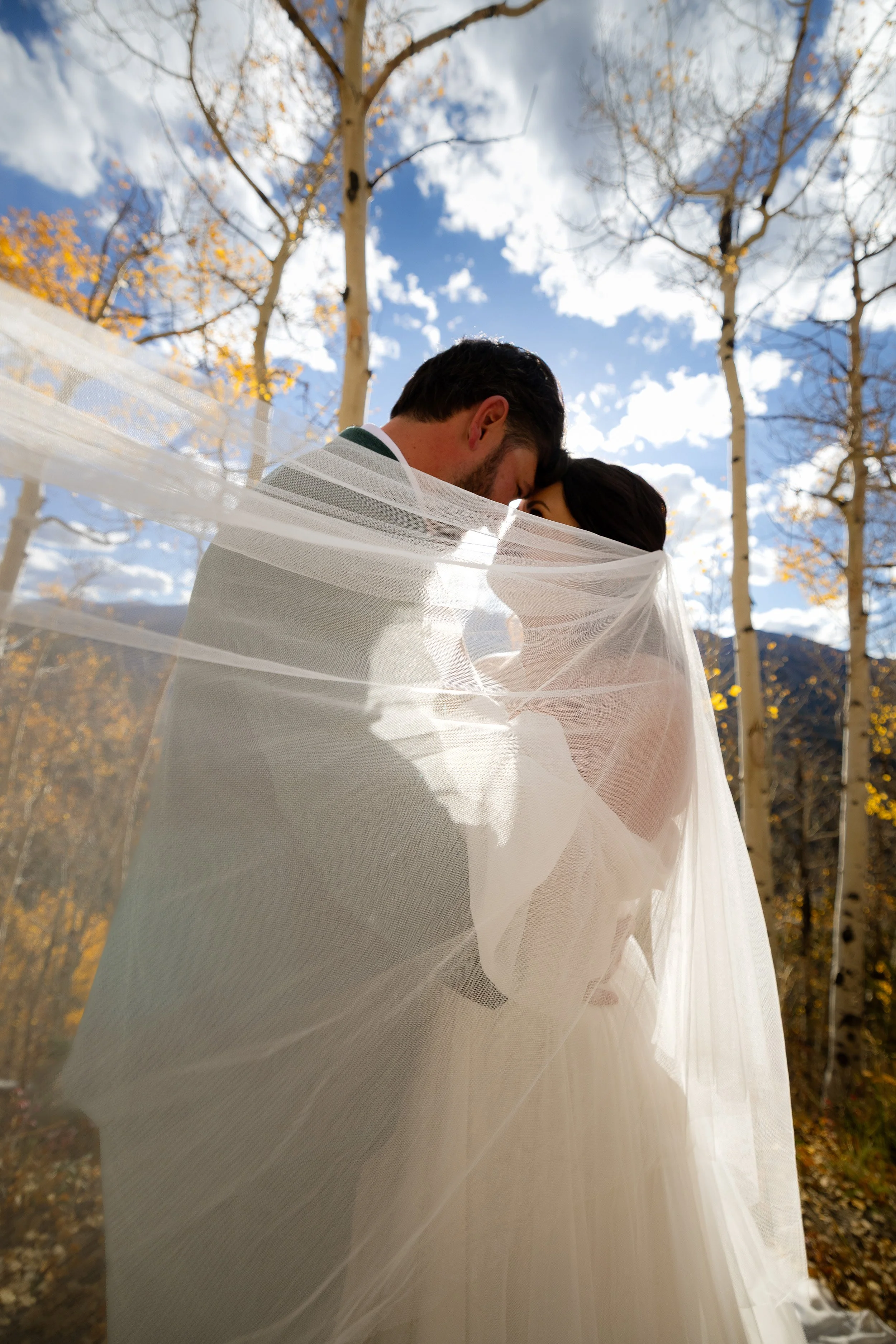 A bride and groom embrace outdoors, wrapped in a sheer veil with autumn trees and blue sky in the background.