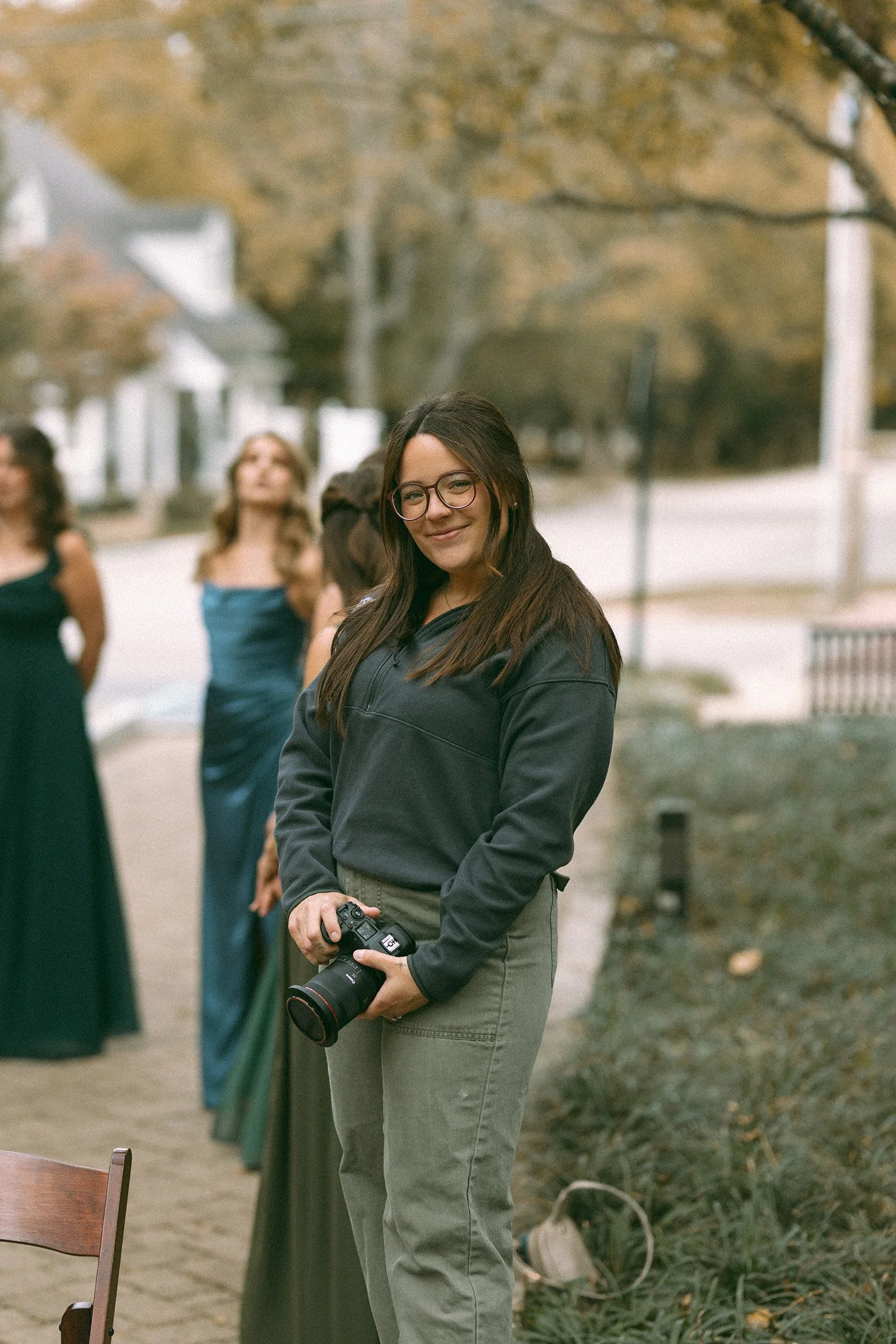 A young woman with glasses and long dark hair holding a camera, smiling, standing outdoors among other women in formal dresses.