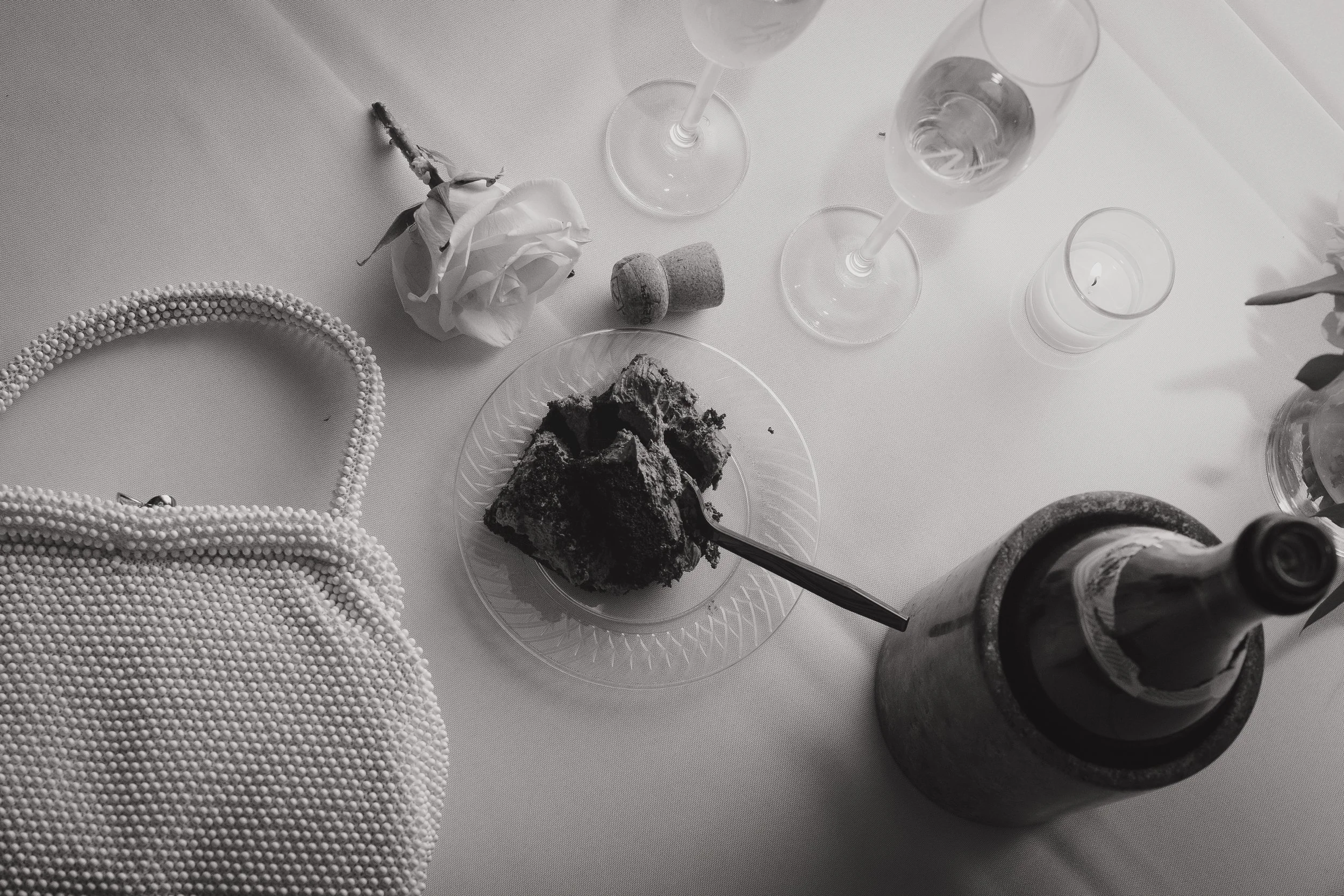 A table with a piece of cake on a glass plate, surrounded by a pearled handbag, a rose, champagne glasses, a bottle in an ice bucket, a candle, and a cork, all in black and white.