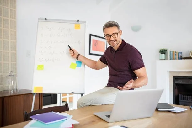 A smiling man with glasses in a maroon shirt and light-colored pants is pointing at a whiteboard in a home office, sitting on a table with a laptop and notebooks.