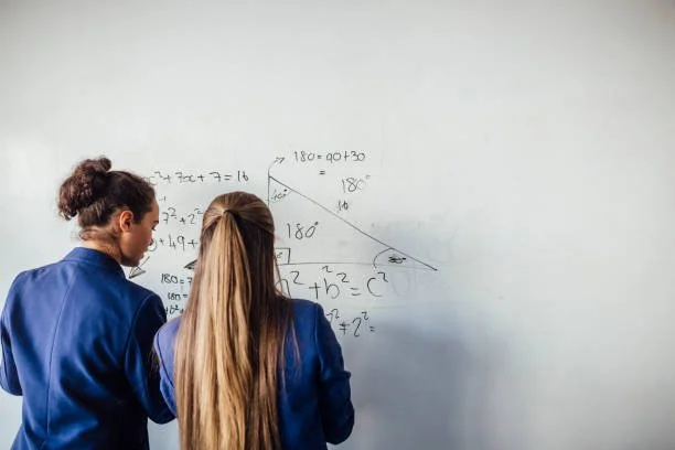 Two students, one with curly hair and one with straight hair, are standing and writing on a classroom whiteboard with basic exercises including addition and a geometry diagram involving a triangle with angles labeled 180 degrees.