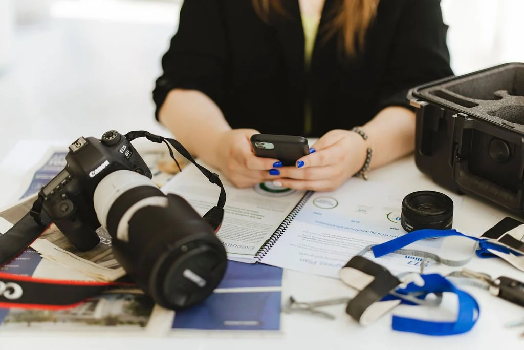 Alyce Peterson sitting at a desk surrounded by camera equipment, documents, and a smartphone.