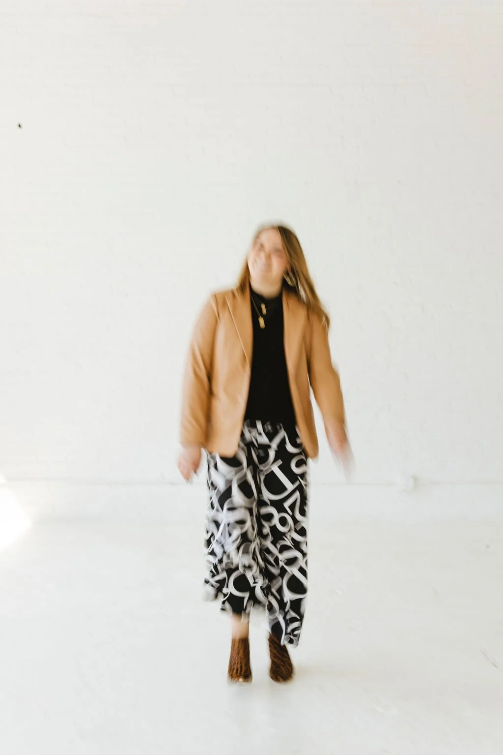 Alyce Peterson wearing leopard print heels, black and white patterned pants, black top, and tan blazer standing against a white wall.